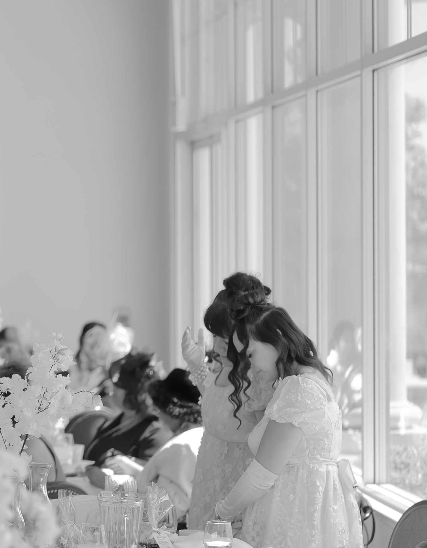 Two women in white dresses share an emotional moment at a wedding reception table near large windows.