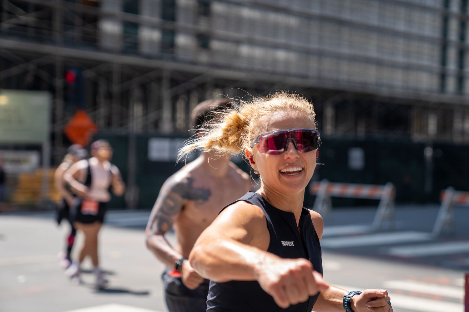 Woman runner in sunglasses smiling during urban race, wearing black tank top and sports watch on sunny day.