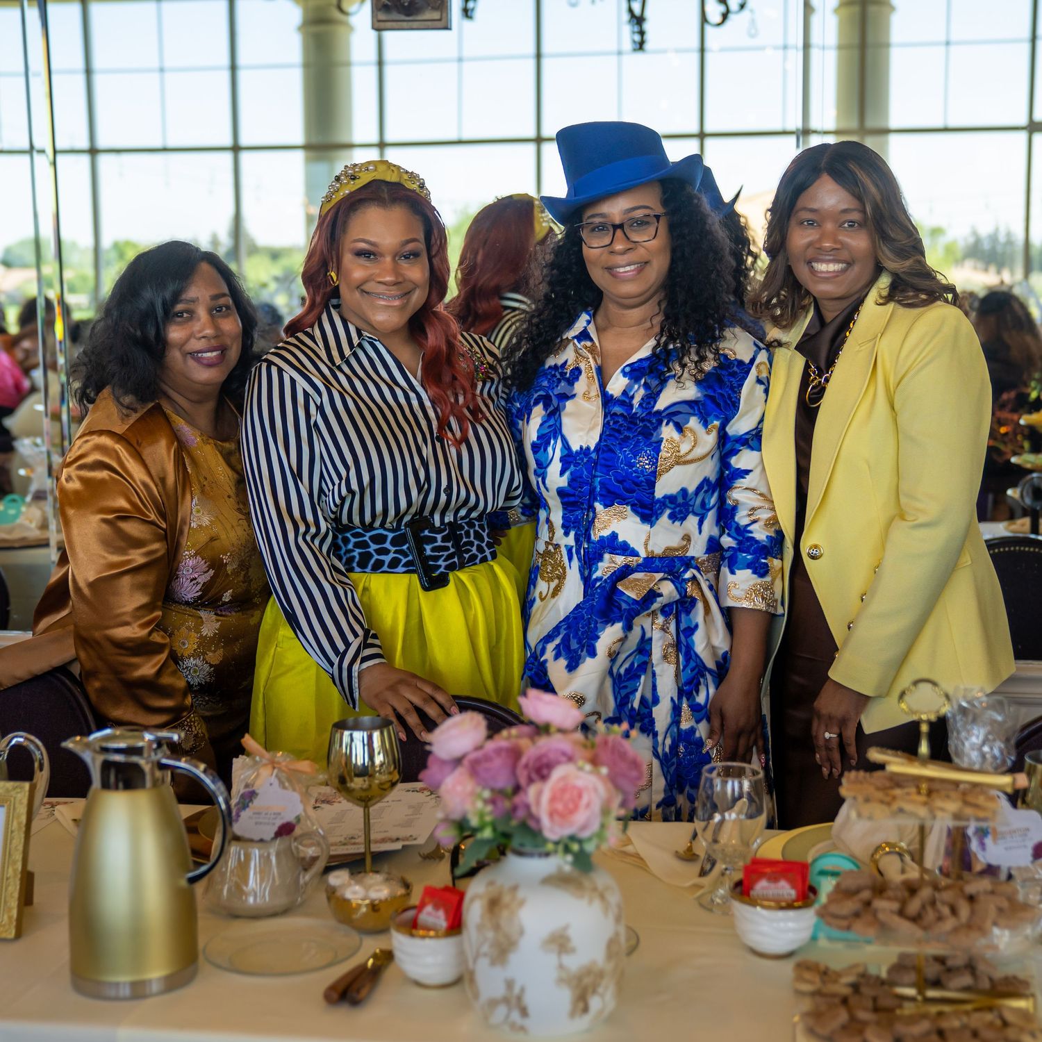 Four elegantly dressed women pose together at a festive table with floral arrangements and tea service.