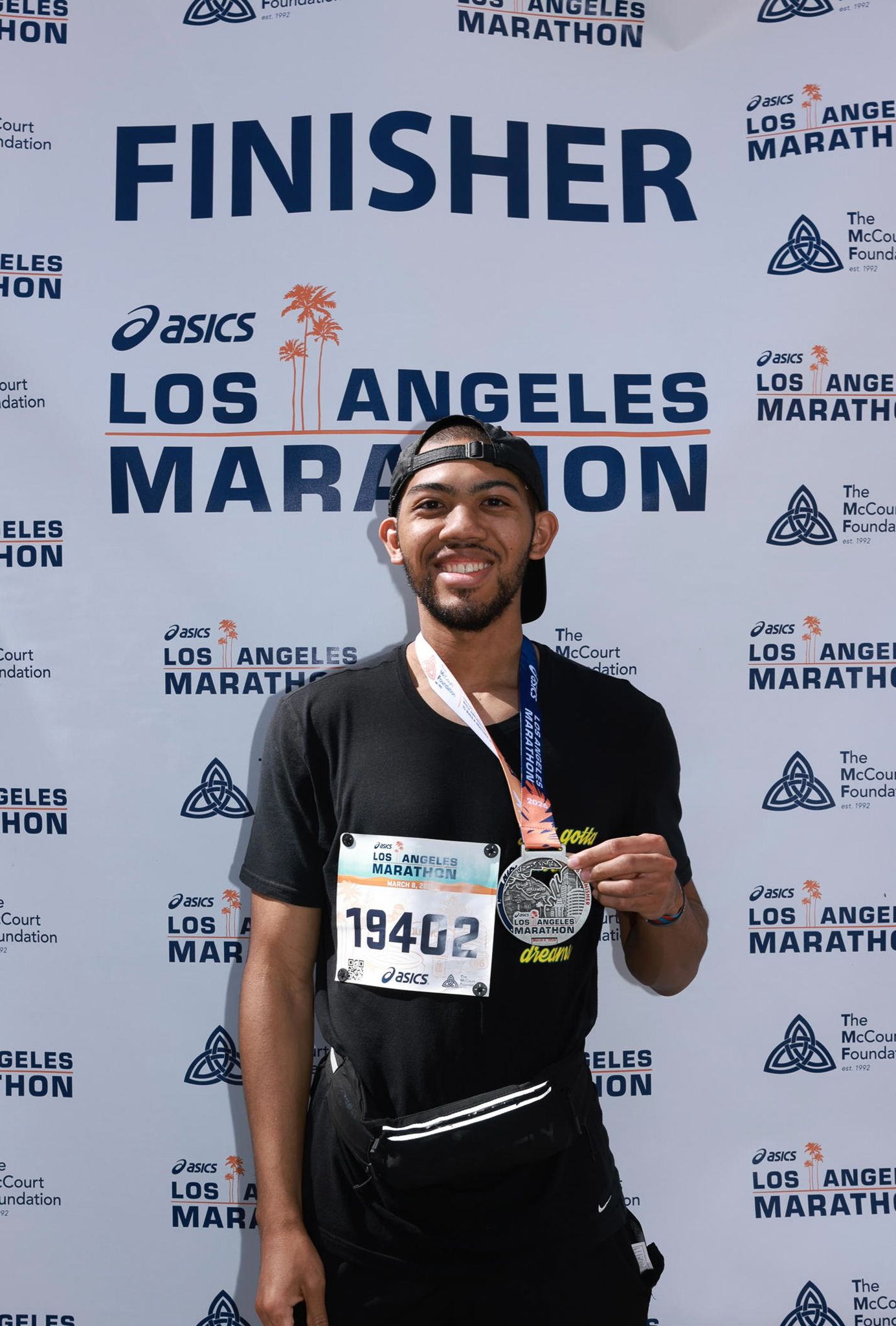 Smiling male runner displays finisher medal at ASICS Los Angeles Marathon finish line backdrop, wearing bib number 19402.