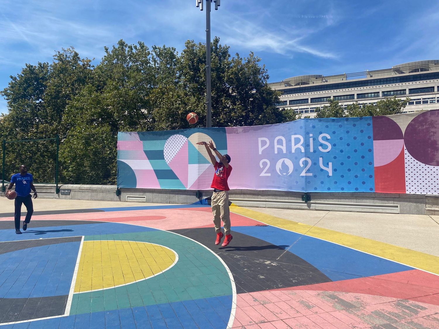 Person playing basketball on a colorful court with Paris 2024 Olympics banner in background on a sunny day.