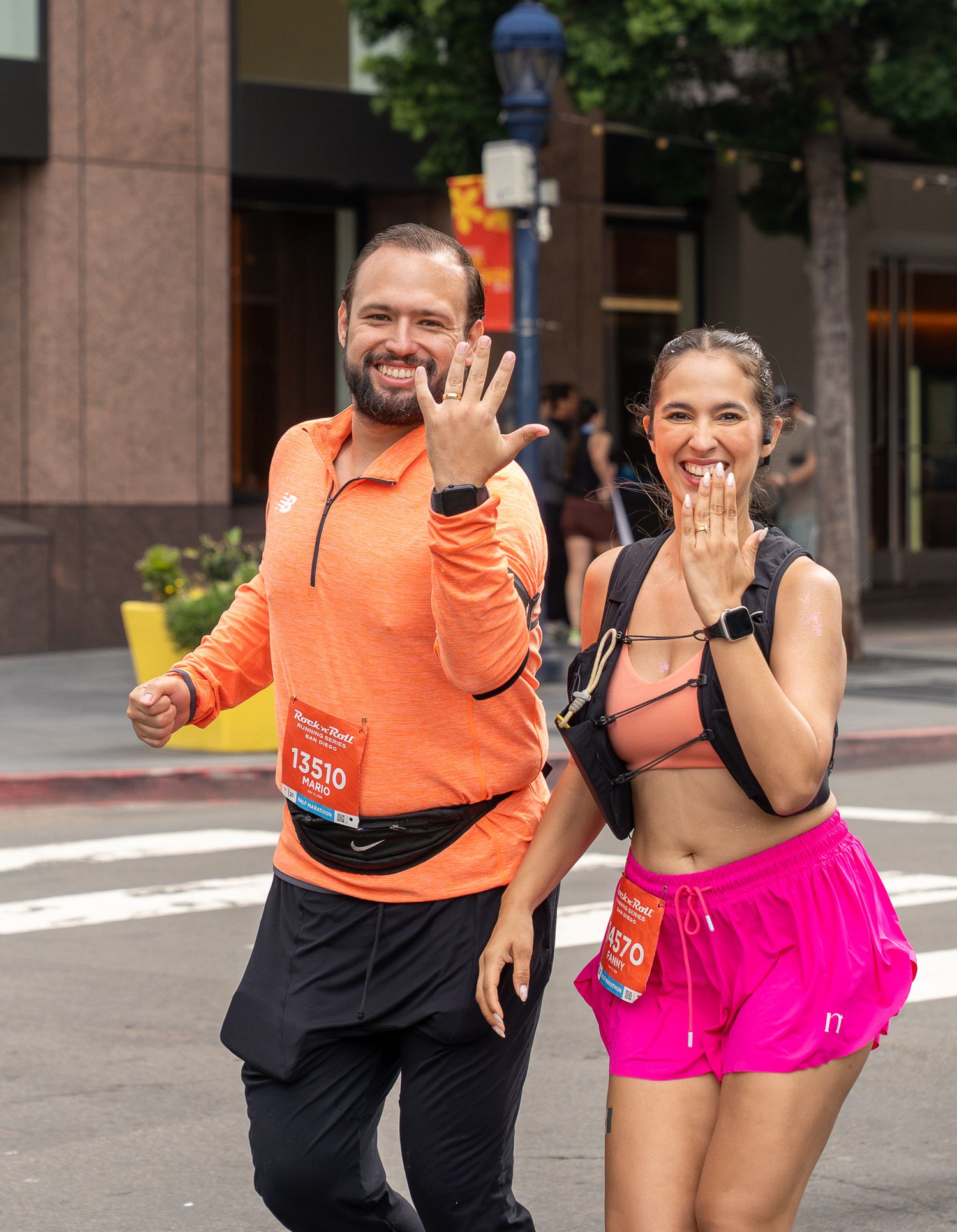 Two runners in race bibs smile and show off rings after a marriage proposal at a city marathon.