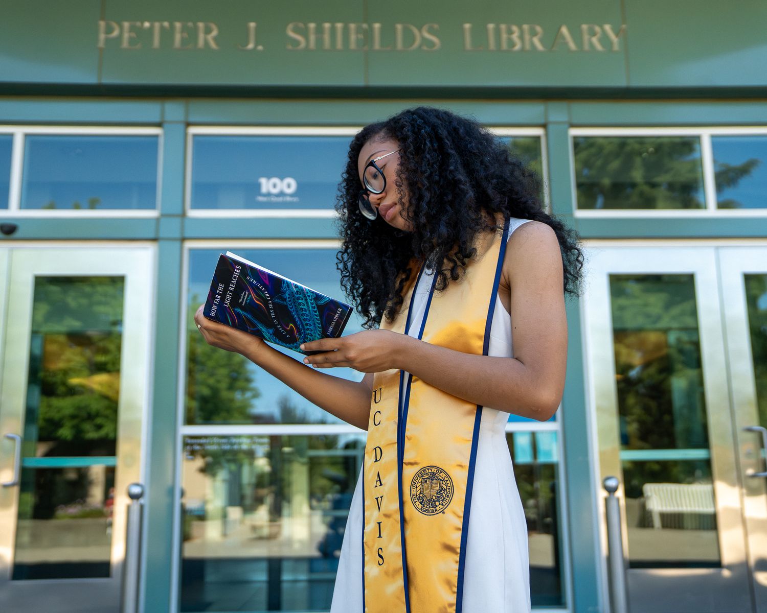 UC Davis graduate in cap and gown reads book outside Peter J. Shields Library on sunny day.