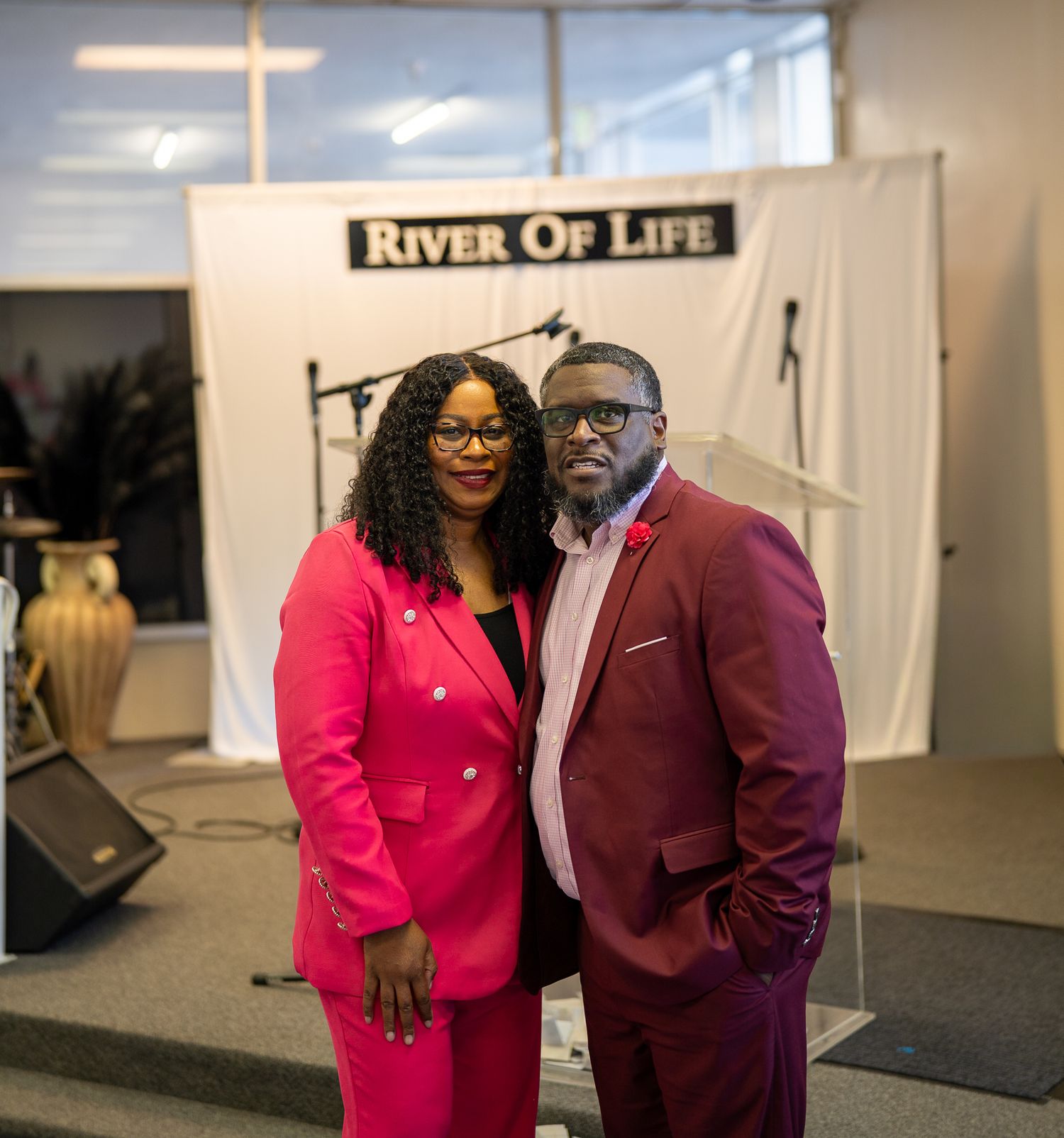 A man and woman in vibrant red and burgundy suits pose together in front of a River of Life church backdrop.