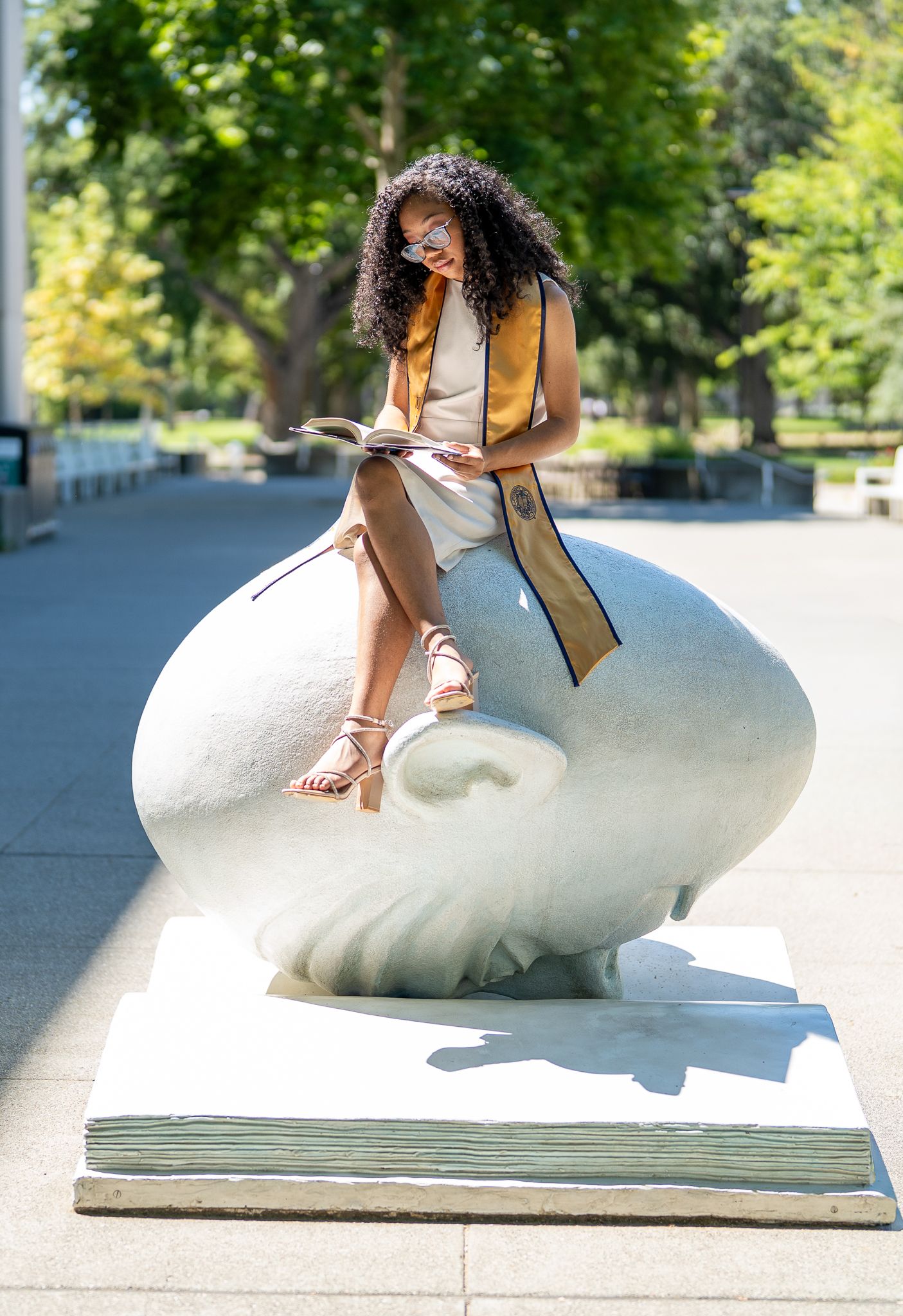 Graduate in cap and gown sash sits on large white sculpture reading a book outdoors on a sunny day.