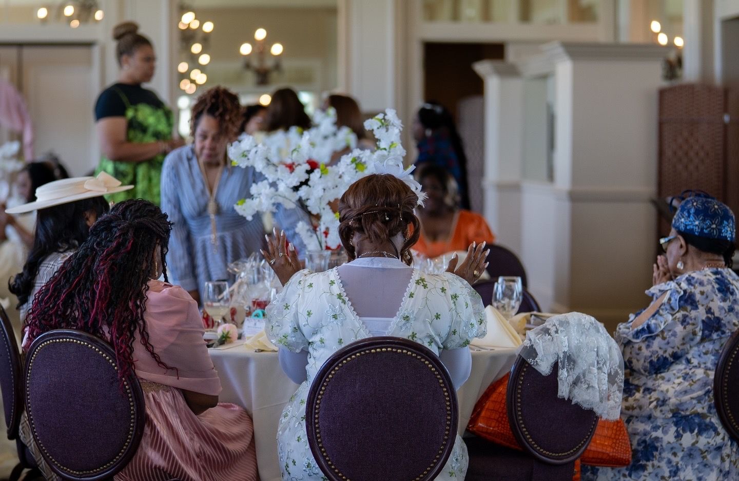 Elegantly dressed women seated at decorated tables during a formal luncheon event in a beautiful banquet hall.