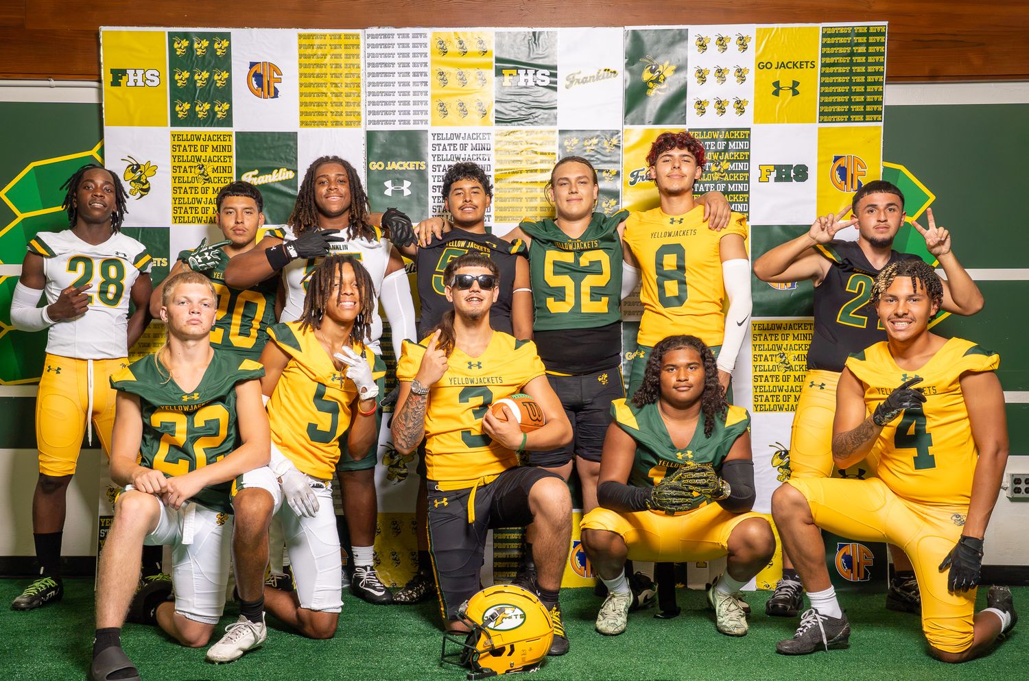 College football team poses in green and gold uniforms with helmets, displaying jerseys in a locker room.