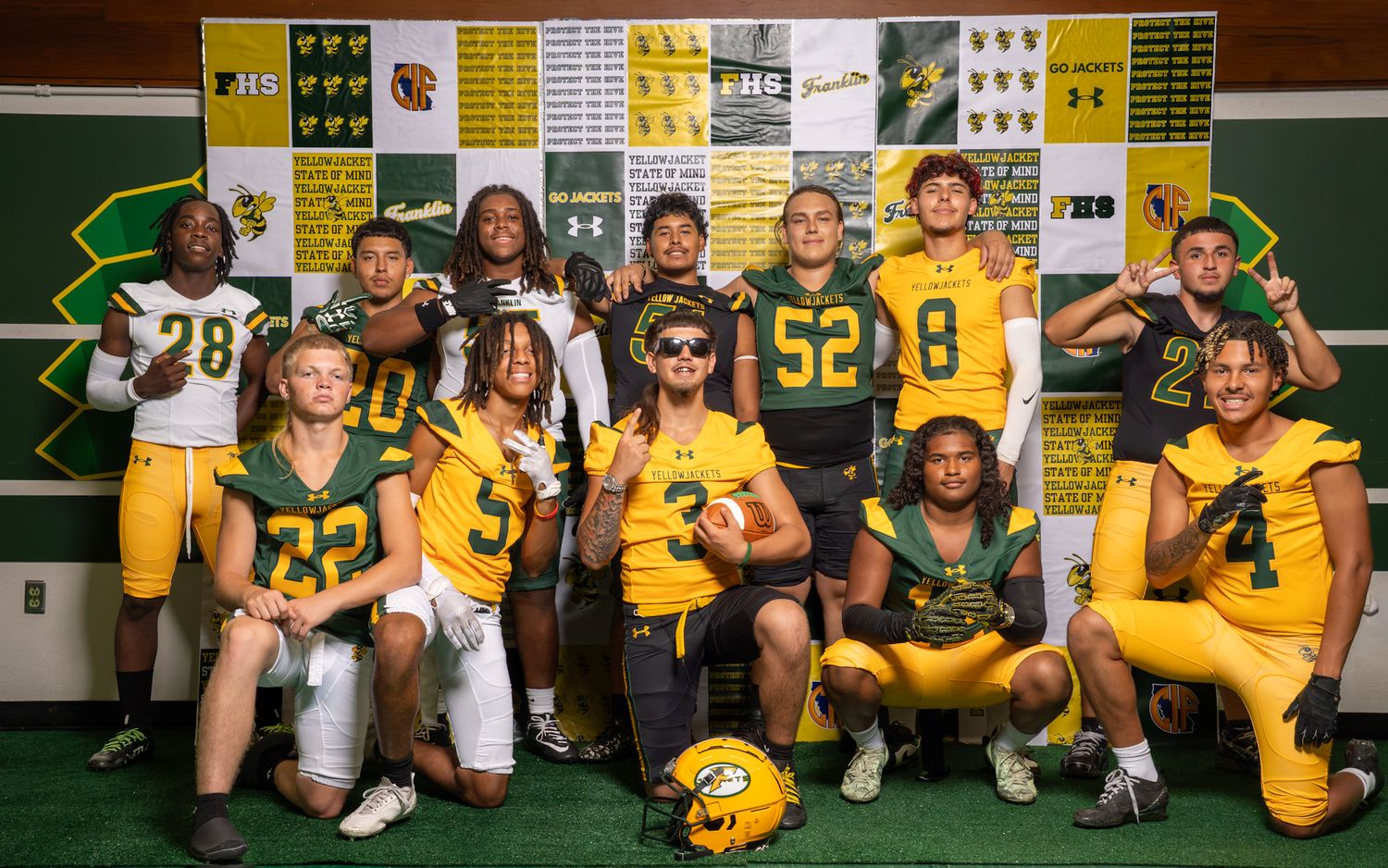 High school football team poses in green and gold uniforms inside a locker room decorated with team banners.