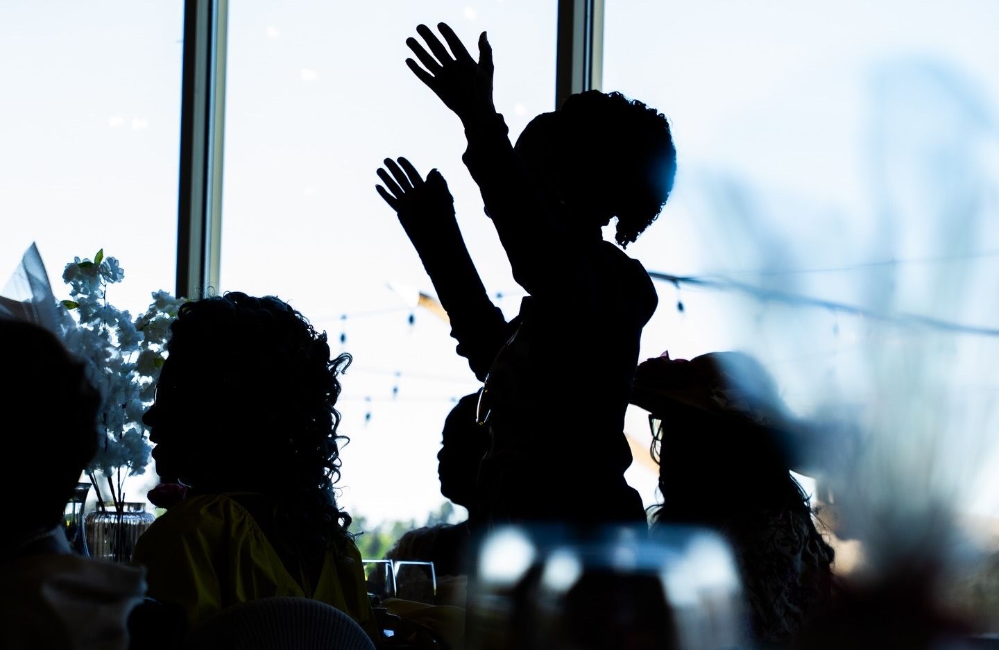 Silhouette of a child raised on shoulders with arms up, surrounded by crowd near bright window.