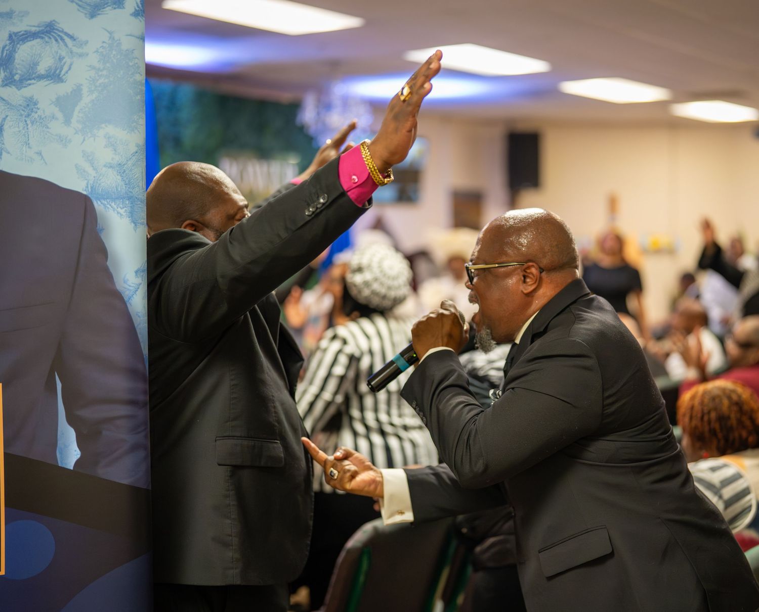 Two men in suits worship expressively at a crowded church service, one raising his hand in praise.