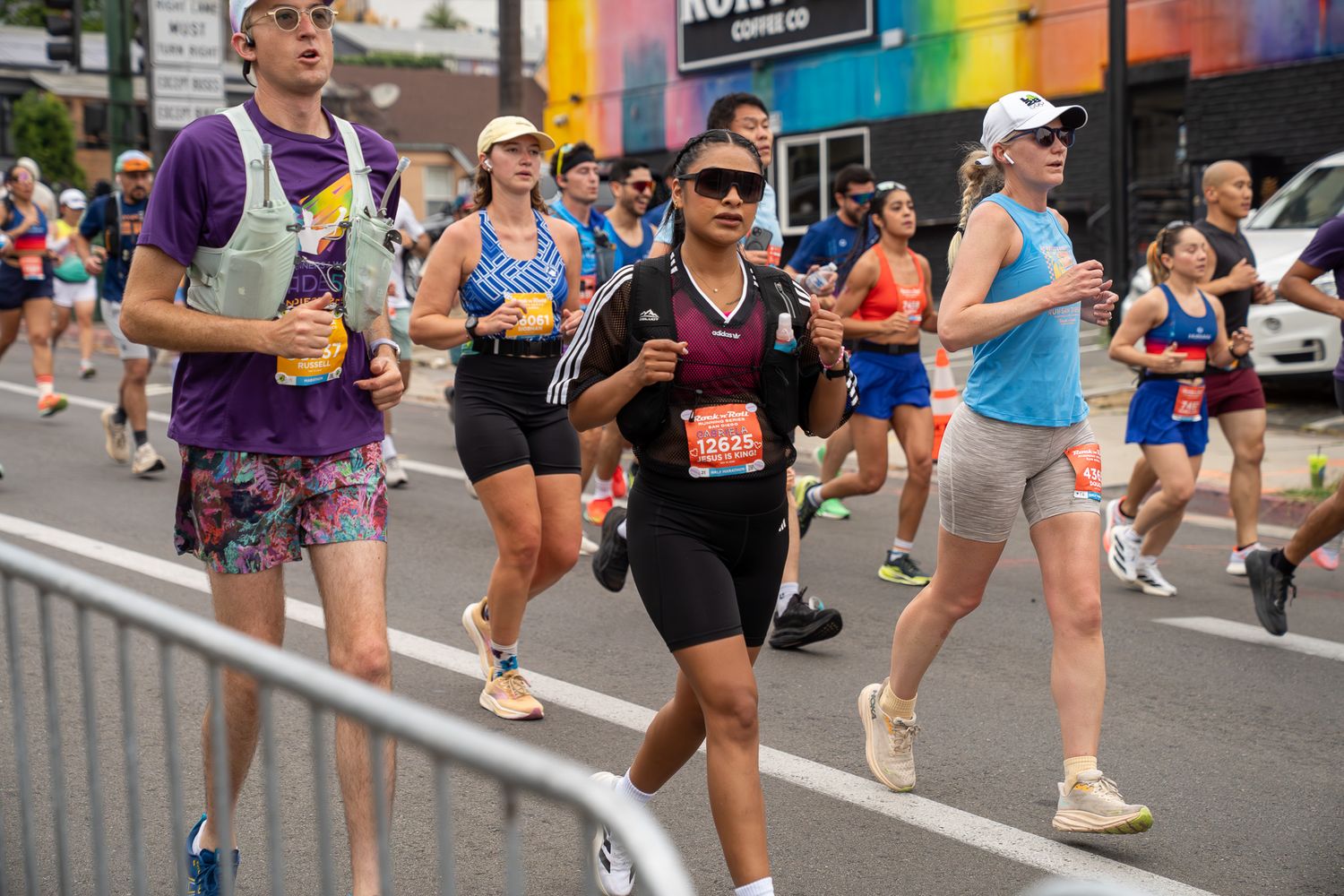 Runners competing in a city marathon race, wearing athletic gear and race bibs, passing storefronts on a sunny day.