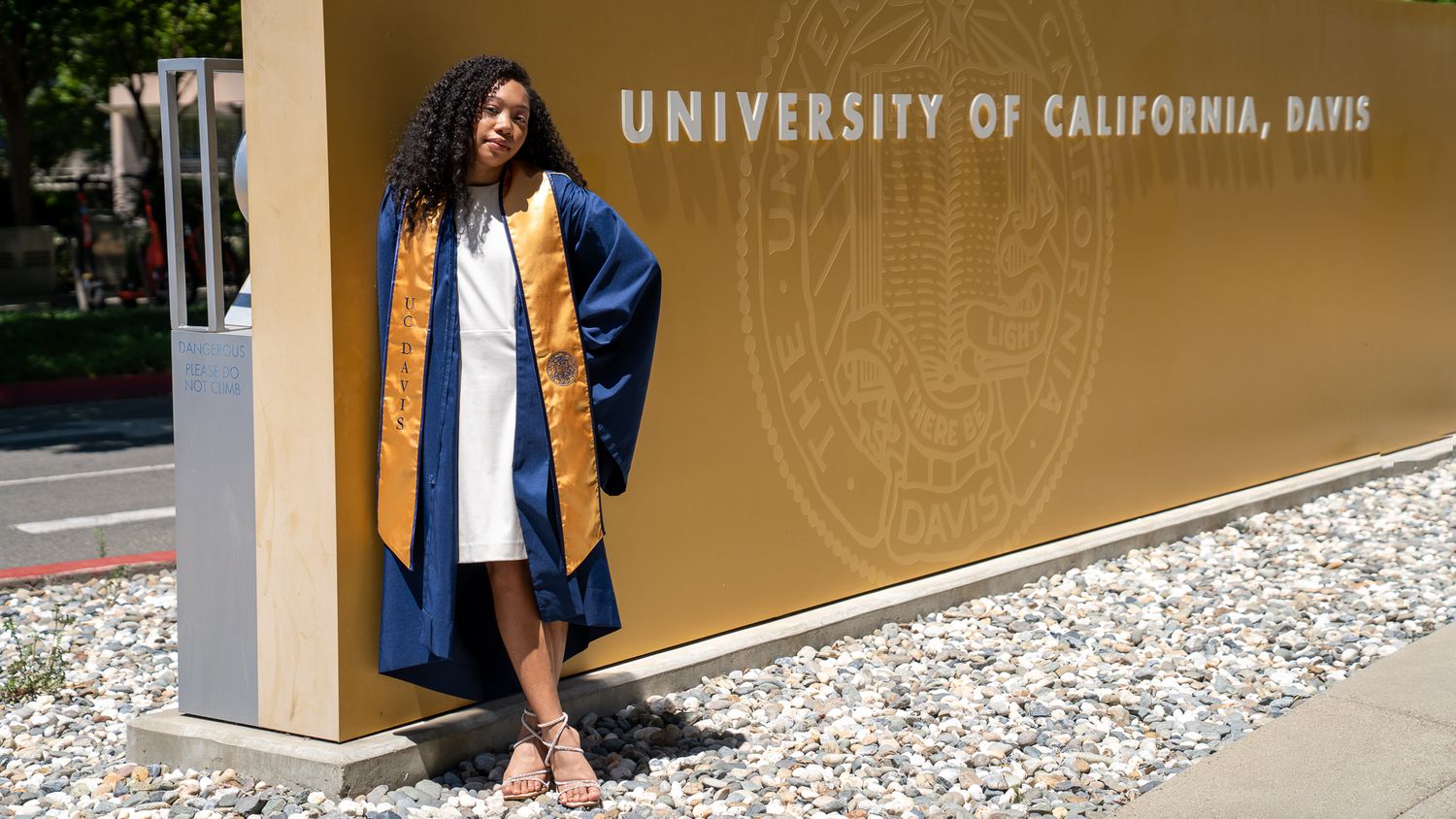 UC Davis graduate in blue cap and gown with gold stole posing by University of California Davis sign.