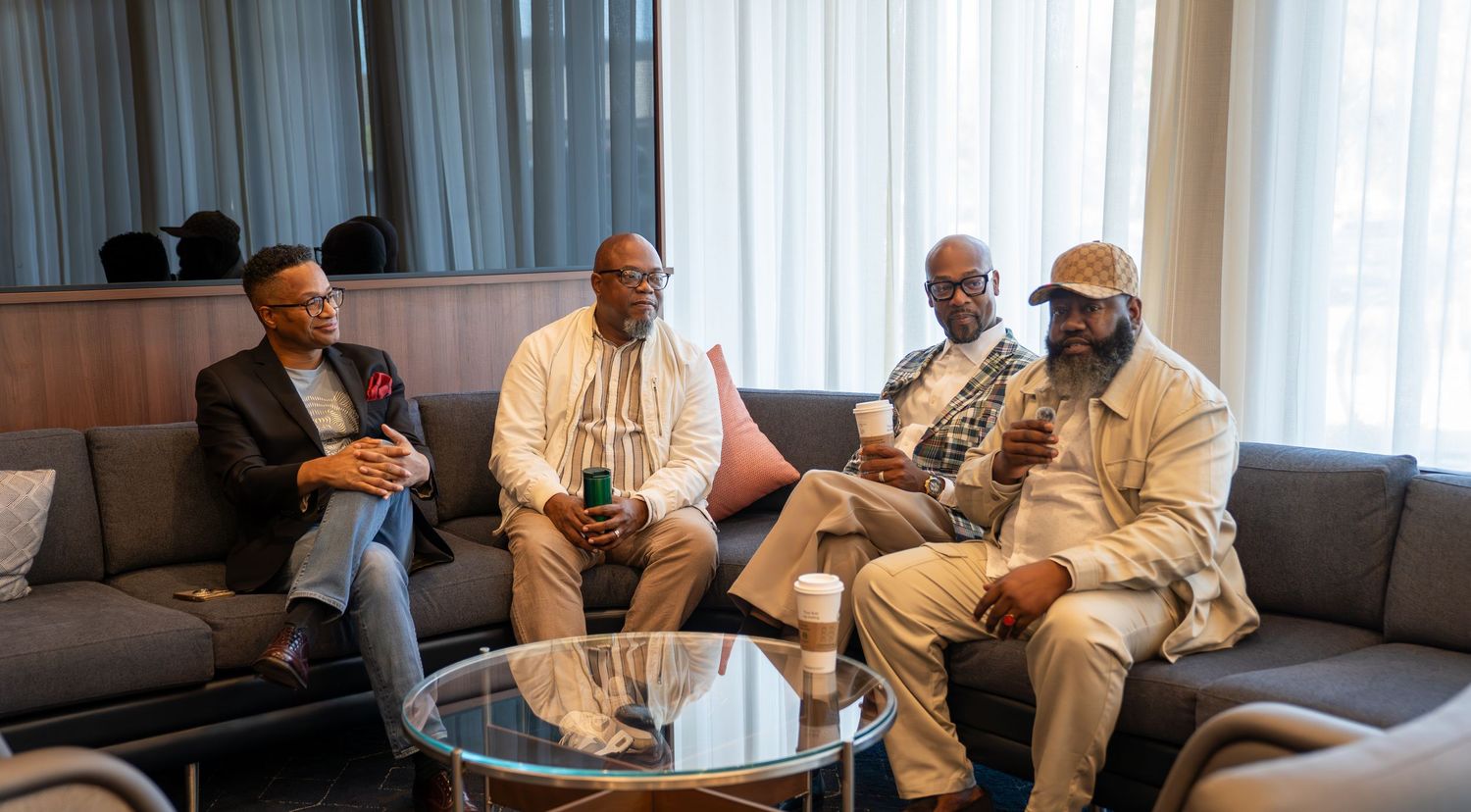 Four Black men sit on gray sofas in a modern lounge, engaged in conversation over coffee and drinks.