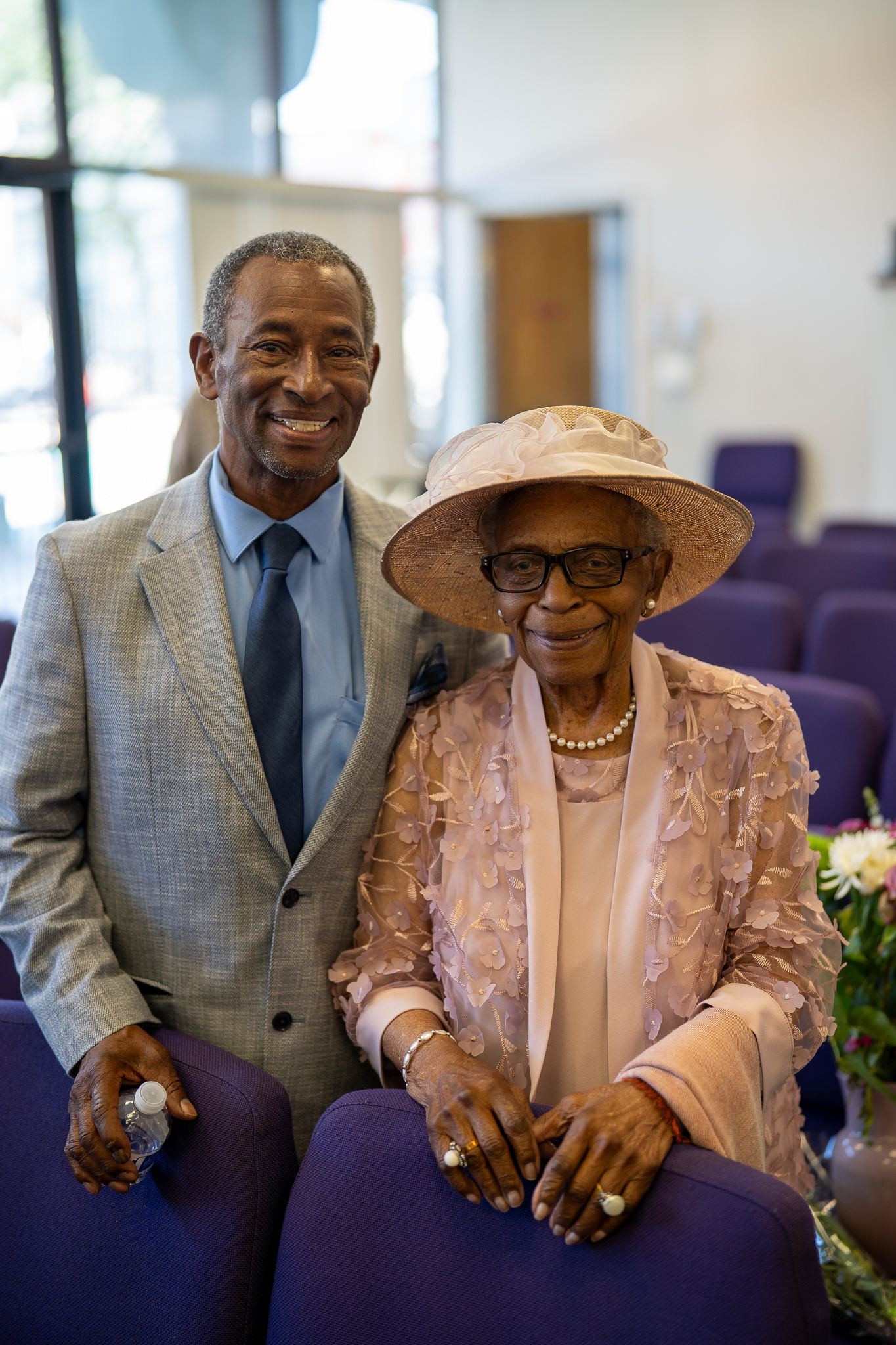 Elderly Black couple dressed elegantly in church, woman wearing pink hat and pearls, man in gray suit smiling warmly.