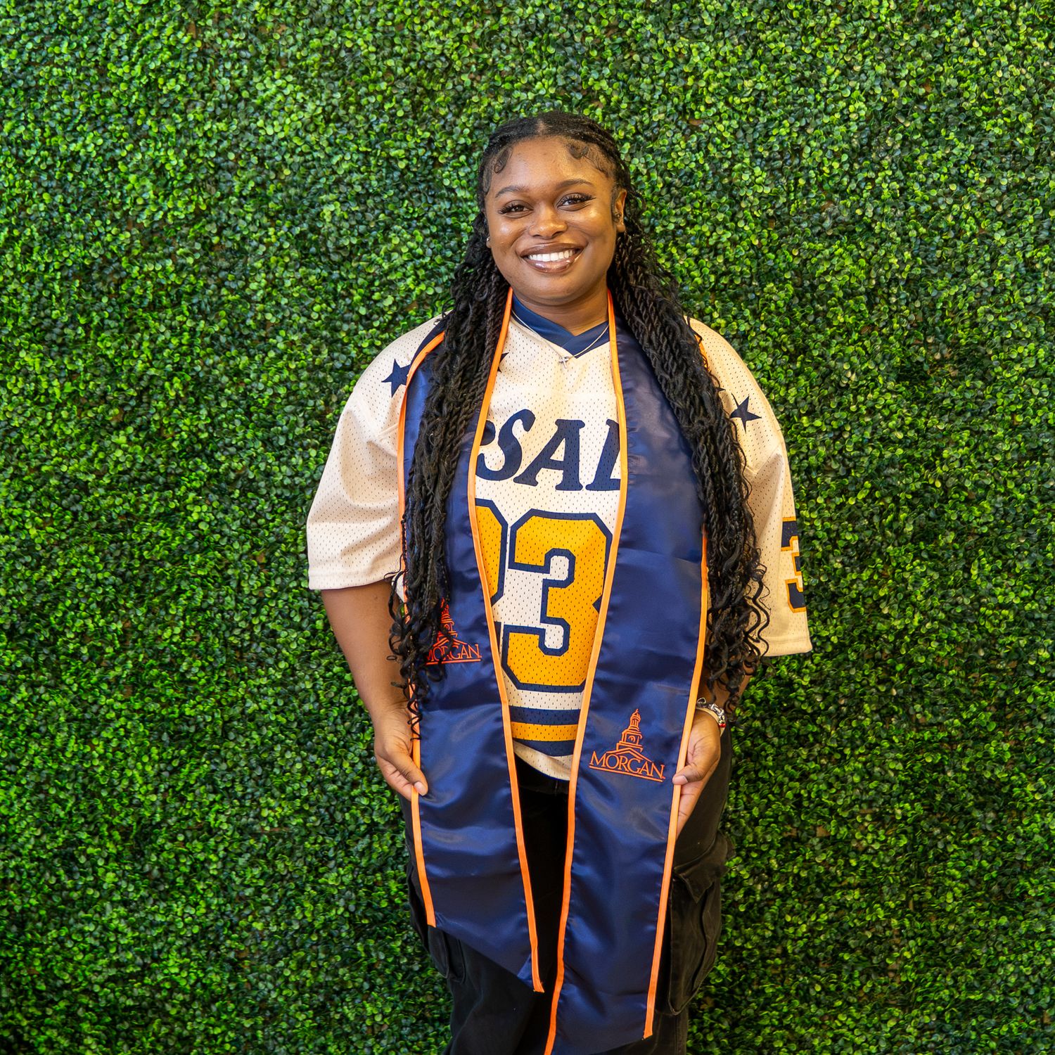 Smiling graduate wearing a CSAL jersey and navy blue graduation stole, posing in front of a green hedge wall.