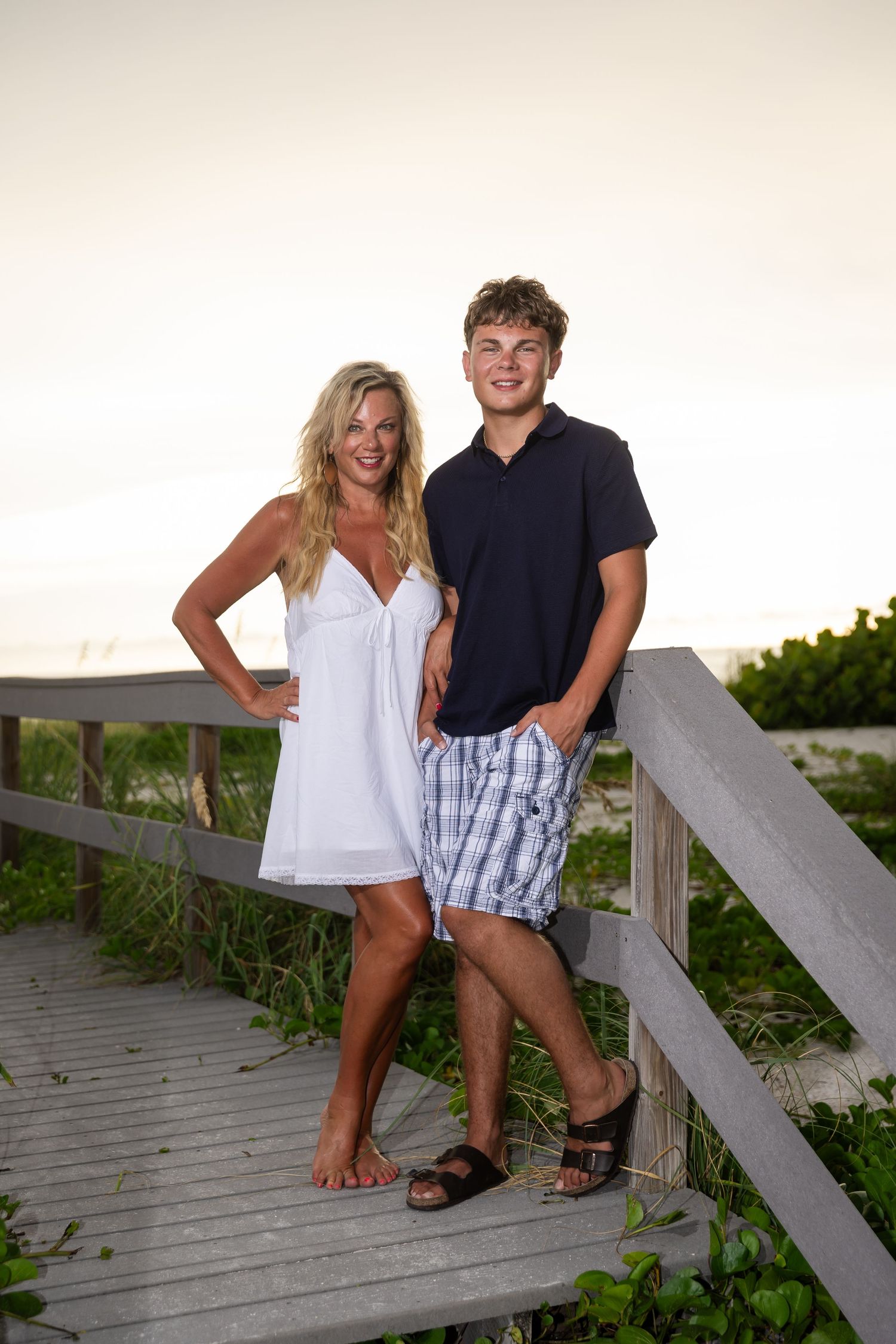 A blonde woman in a white dress and a young man in a navy polo pose together on a beach boardwalk at sunset.