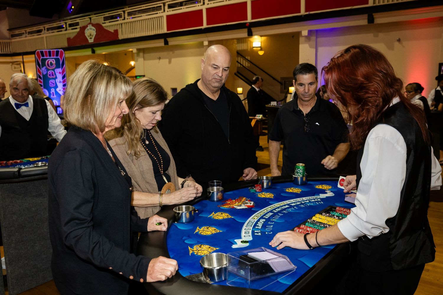 Guests gather around a blue blackjack table at a casino night fundraiser event in a large hall.