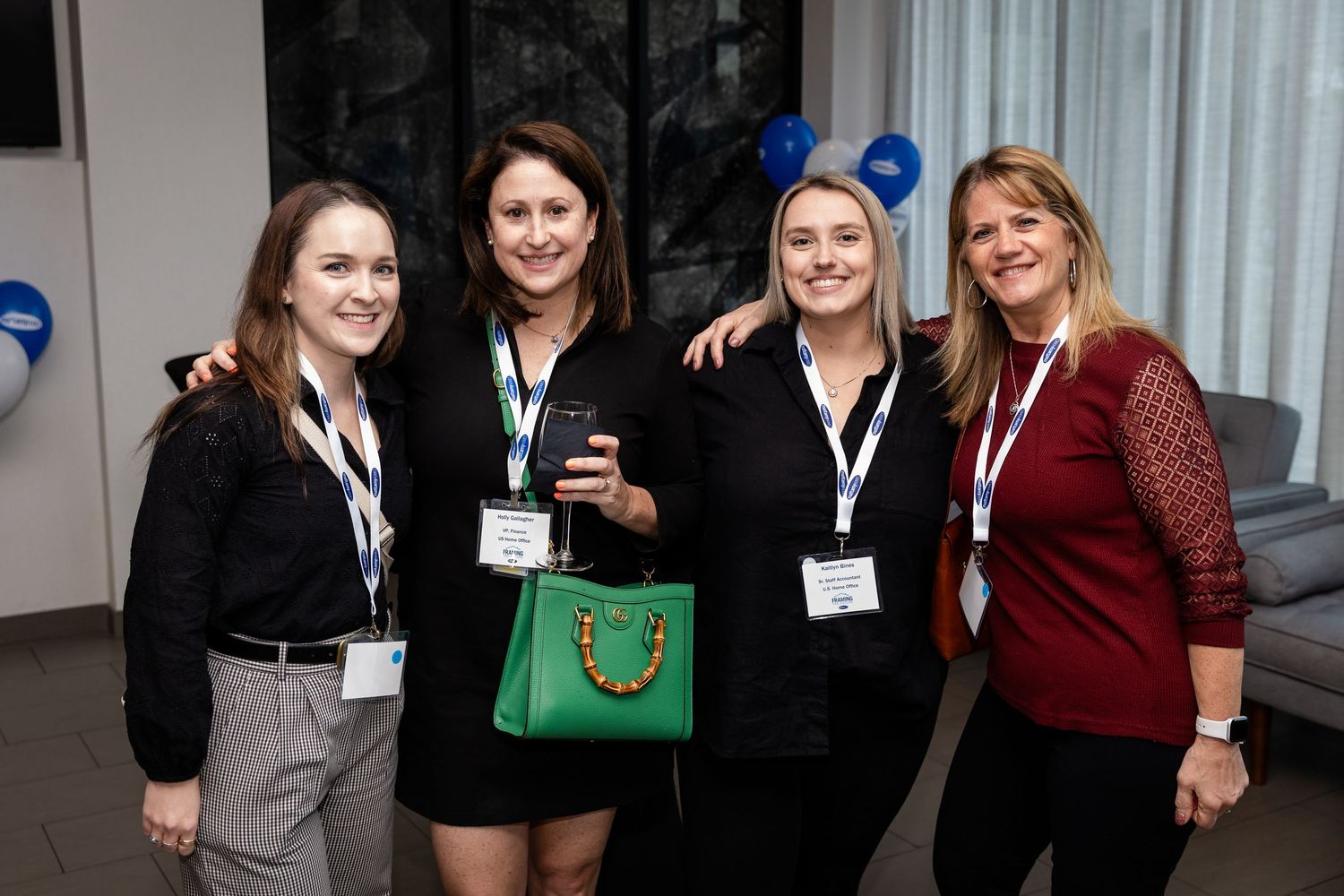Four women smiling and posing together at a professional networking event, wearing lanyards and name badges.