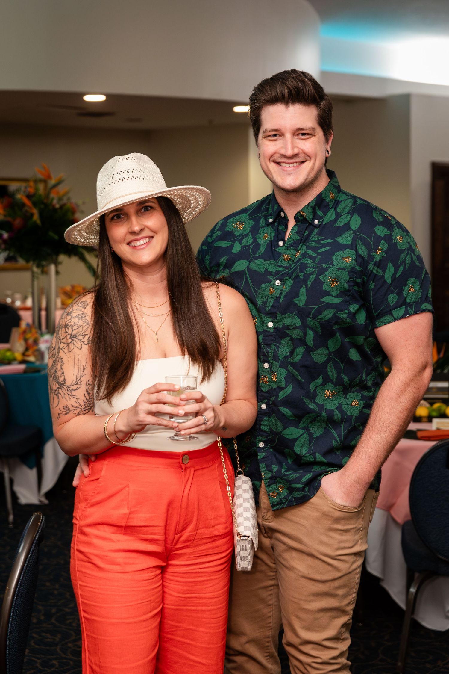 A smiling couple poses at an indoor event, woman in white top, orange pants, and straw hat, man in floral shirt.