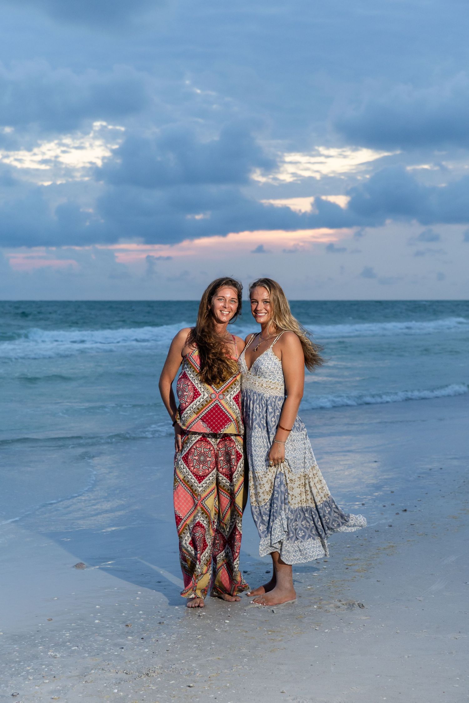 Two women in boho dresses posing together on a white sand beach during a beautiful sunset.