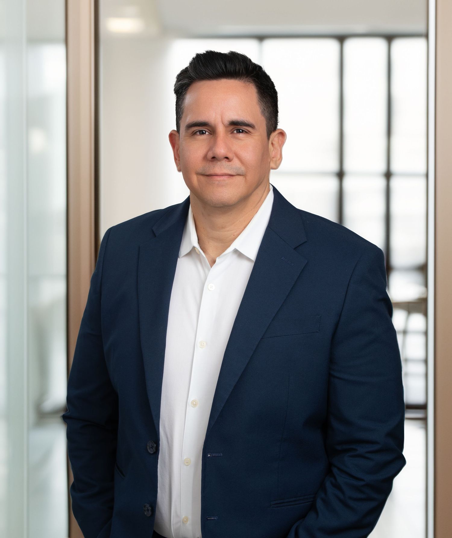 Professional man in navy blazer and white shirt poses confidently in a modern office setting.