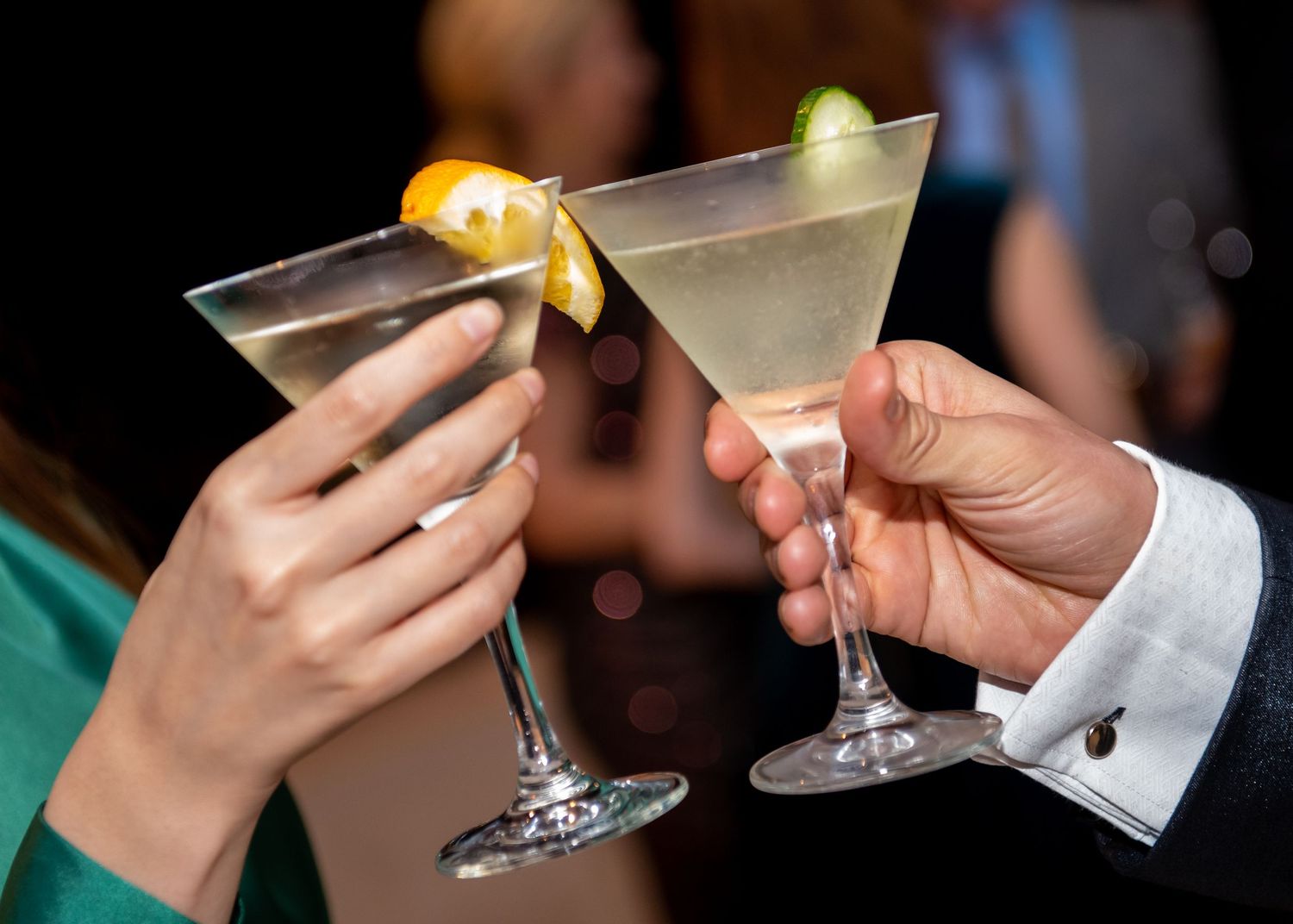 Two hands clinking martini glasses garnished with citrus at a social gathering.