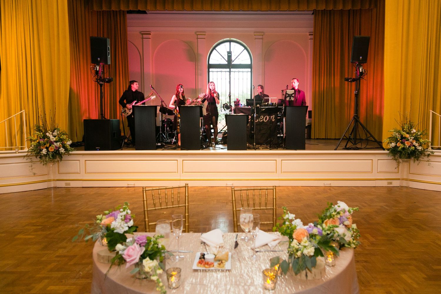 Live wedding band performing on stage in elegant golden ballroom with floral centerpiece in foreground.