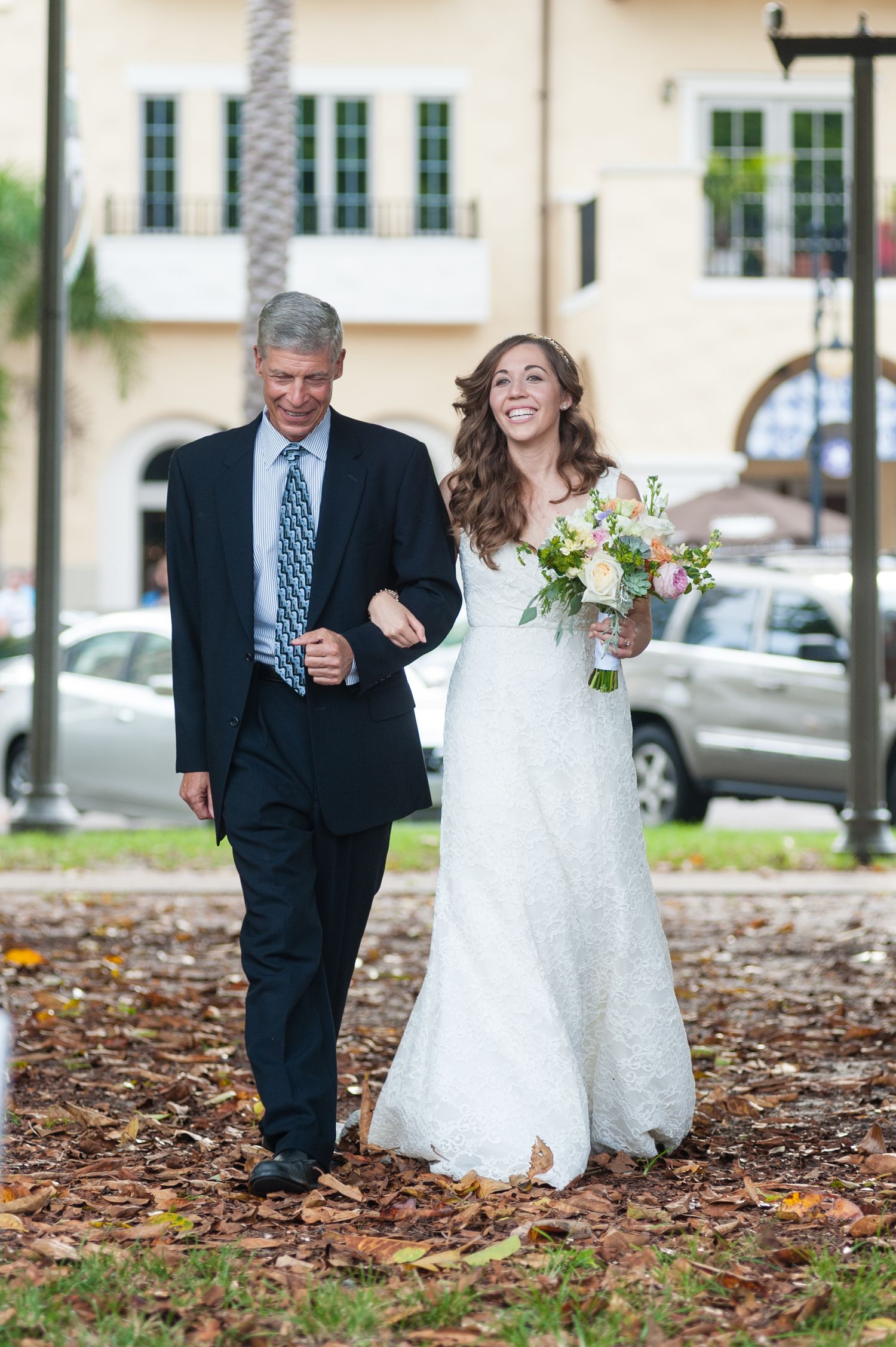 Smiling bride in lace wedding dress walks arm-in-arm with father holding bouquet of pastel flowers.