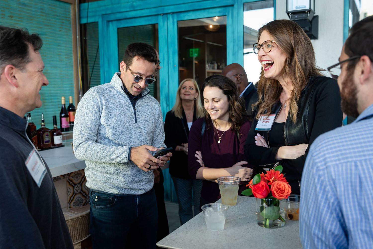 Group of professionals networking at a casual indoor bar event, laughing and looking at a smartphone.