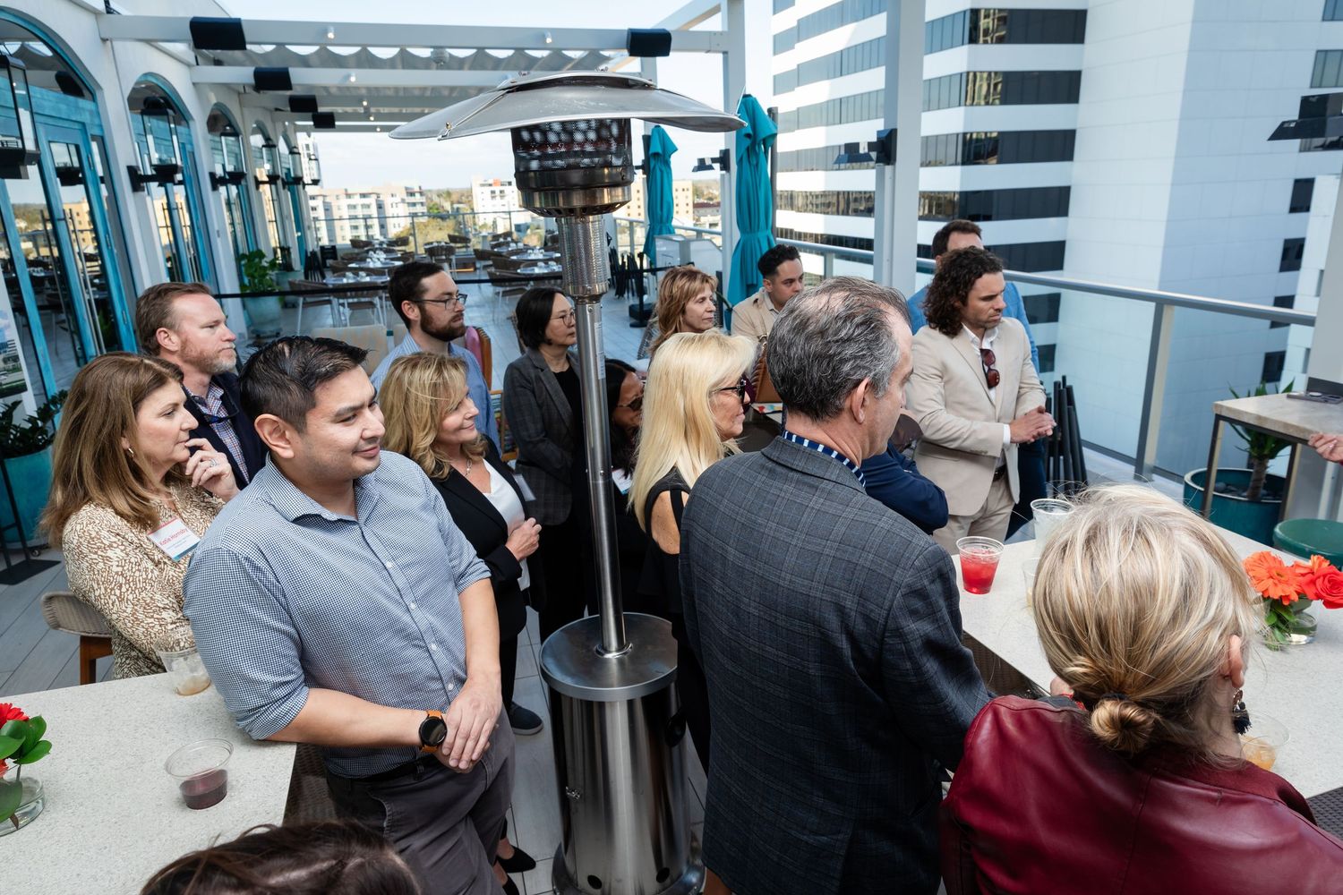 Professionals networking at an outdoor rooftop event with patio heaters and city buildings in the background.