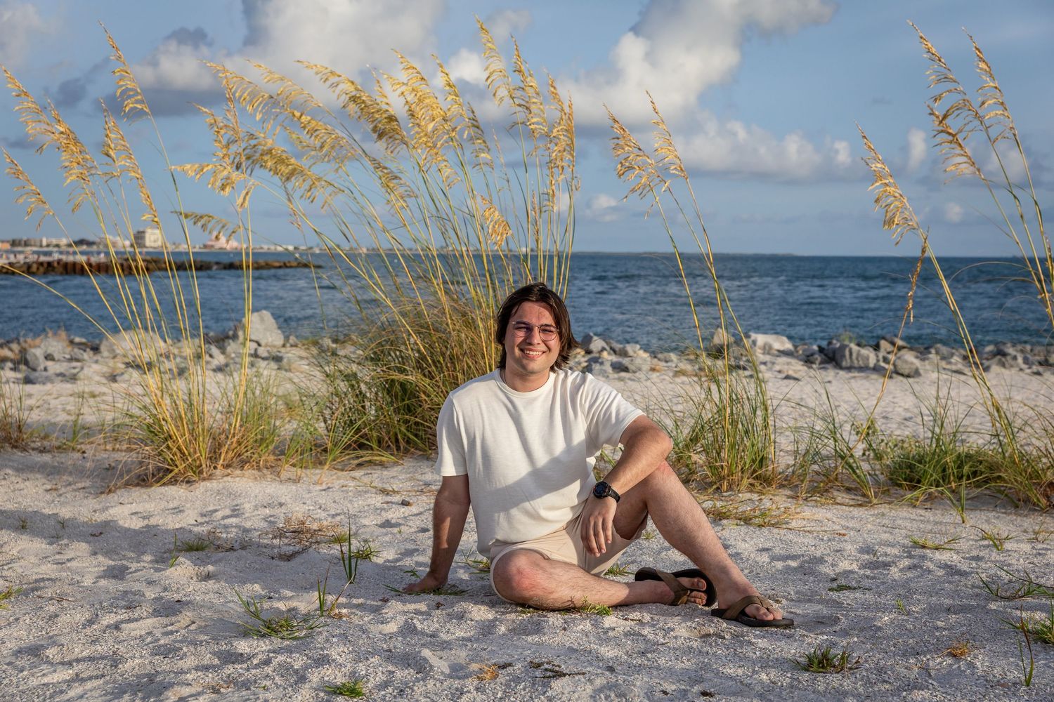 Woman sitting on sandy beach with sea oats, ocean, and pier in background on a sunny day.