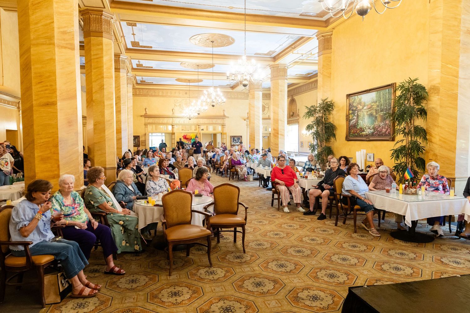 Elderly guests gathered in an elegant hotel ballroom with ornate columns for a large group event or senior gathering.