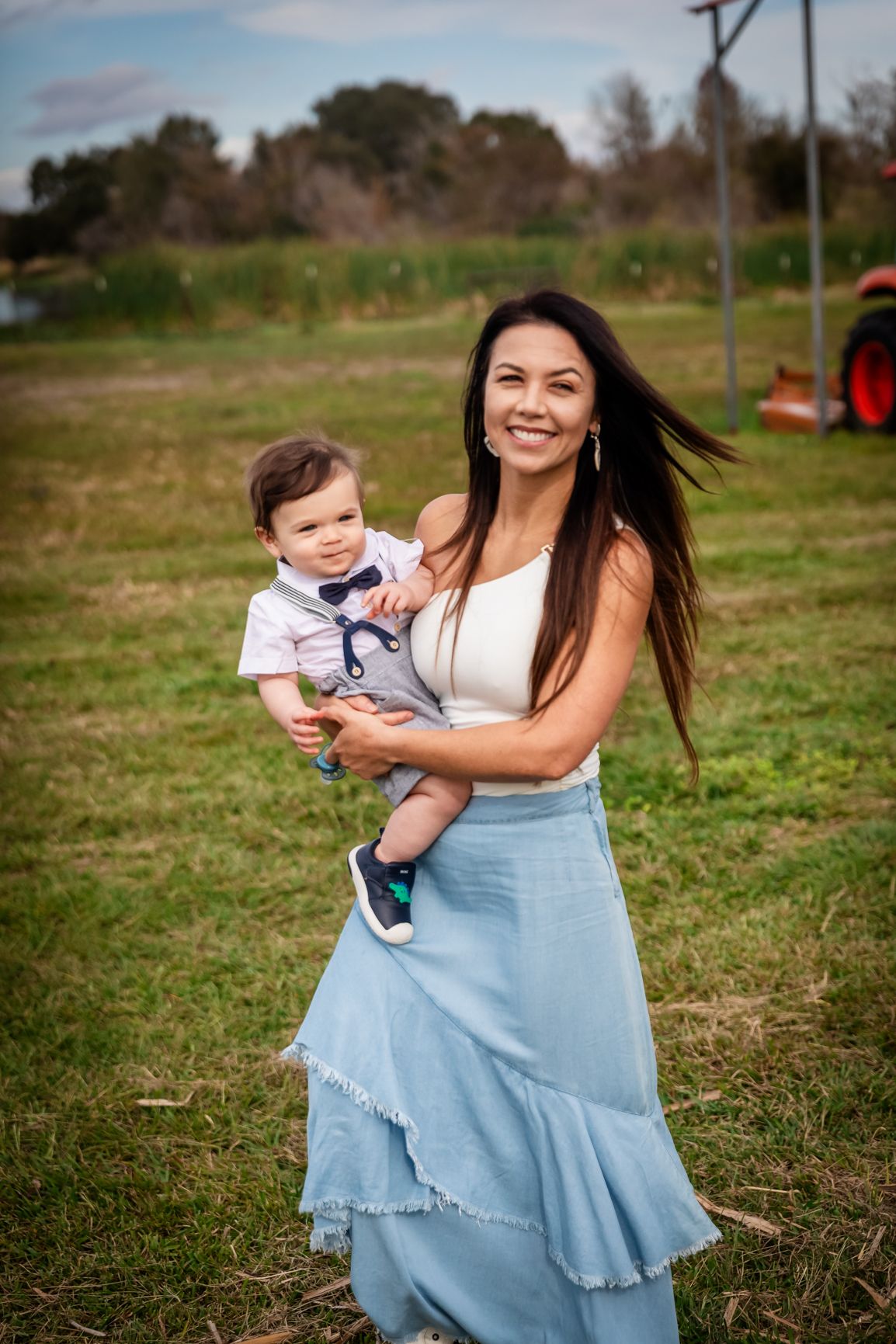 A smiling dark-haired mother in a blue skirt holds her baby boy while standing in an open farm field.