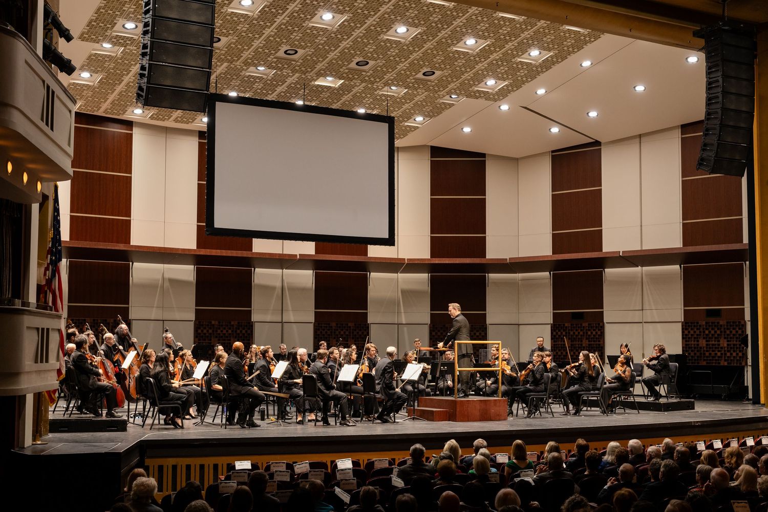 Full symphony orchestra performing on a large concert hall stage before a packed seated audience.