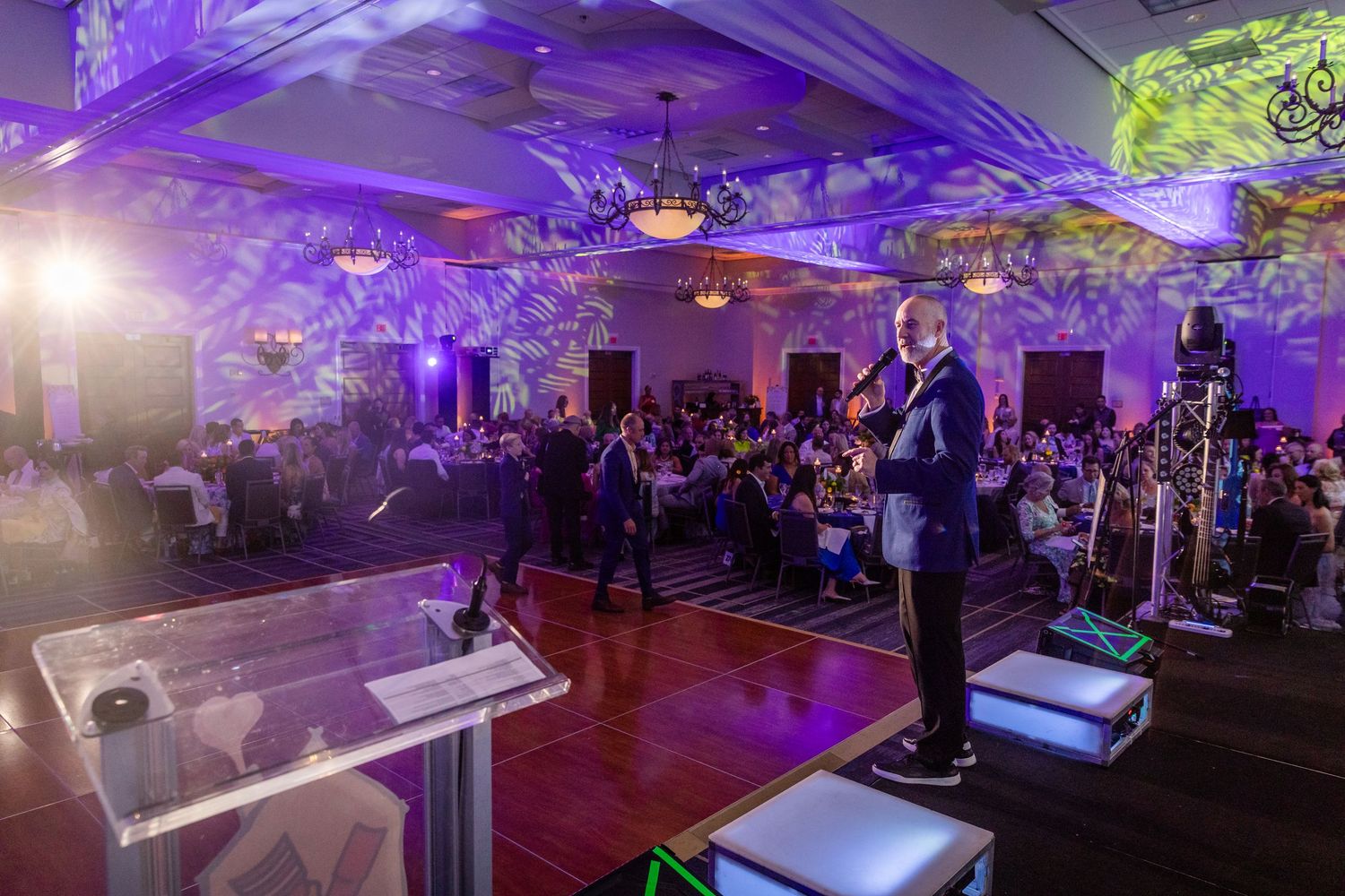 Speaker addresses a large audience in a beautifully lit ballroom with purple lighting.