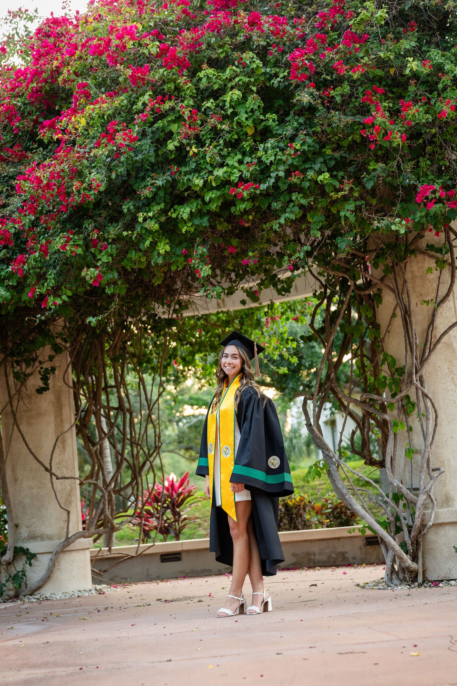 A smiling graduate in black cap and gown with yellow sash poses under a floral arch of vibrant pink bougainvillea.