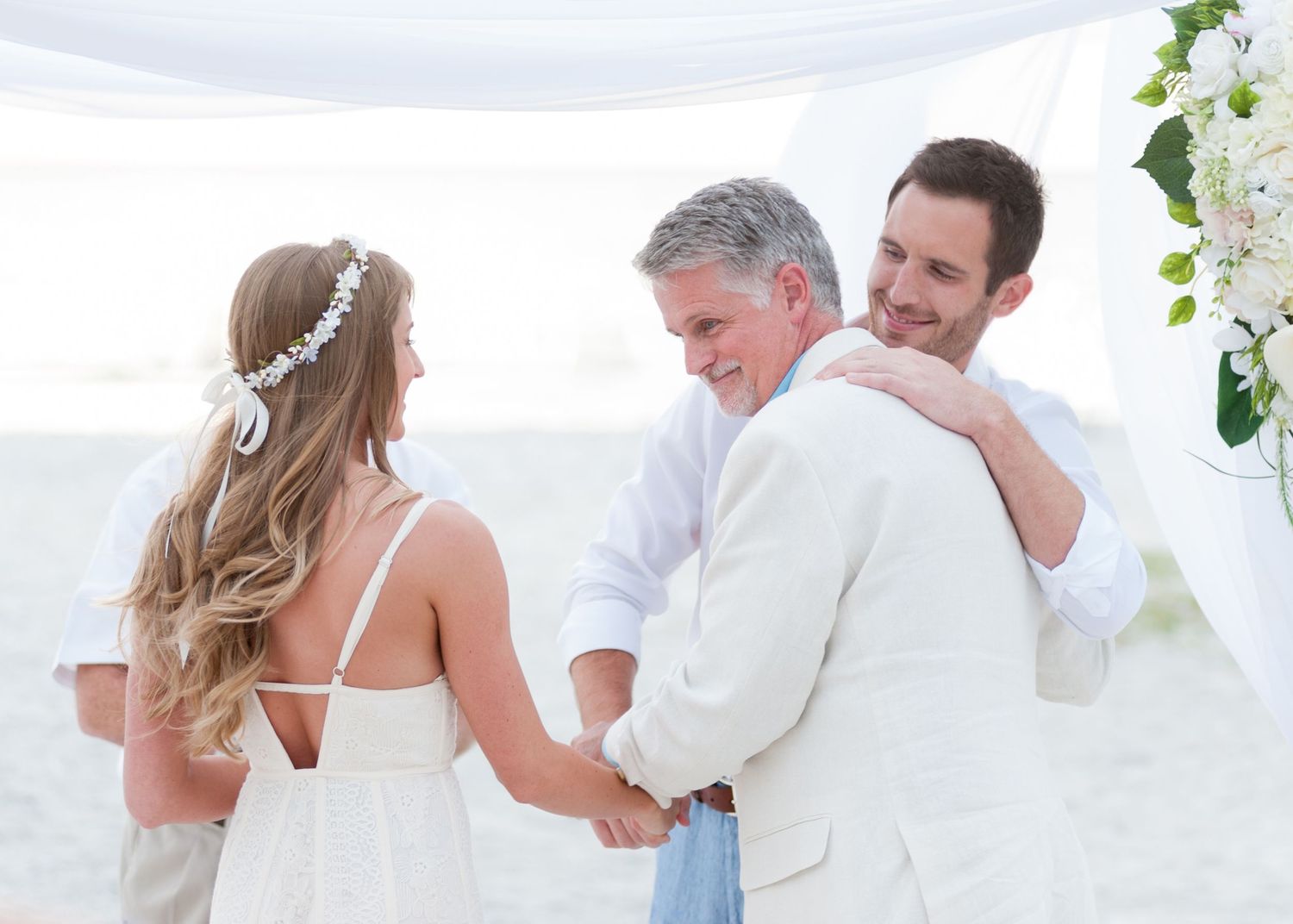 Couple holding hands at beach wedding ceremony with officiant smiling under white canopy with floral arch.