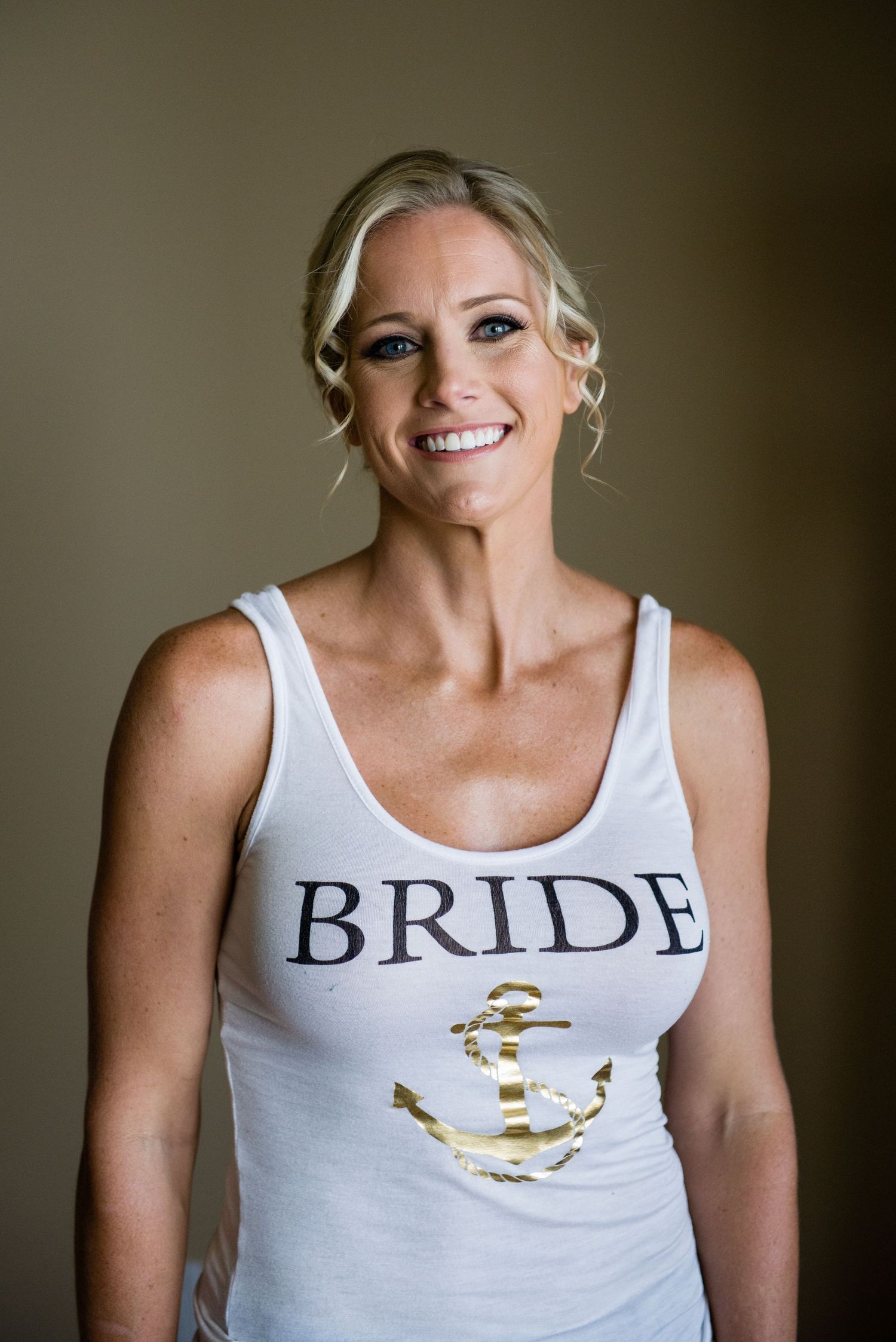 Smiling bride wearing a white tank top with BRIDE text and gold anchor graphic, with elegant updo hairstyle.