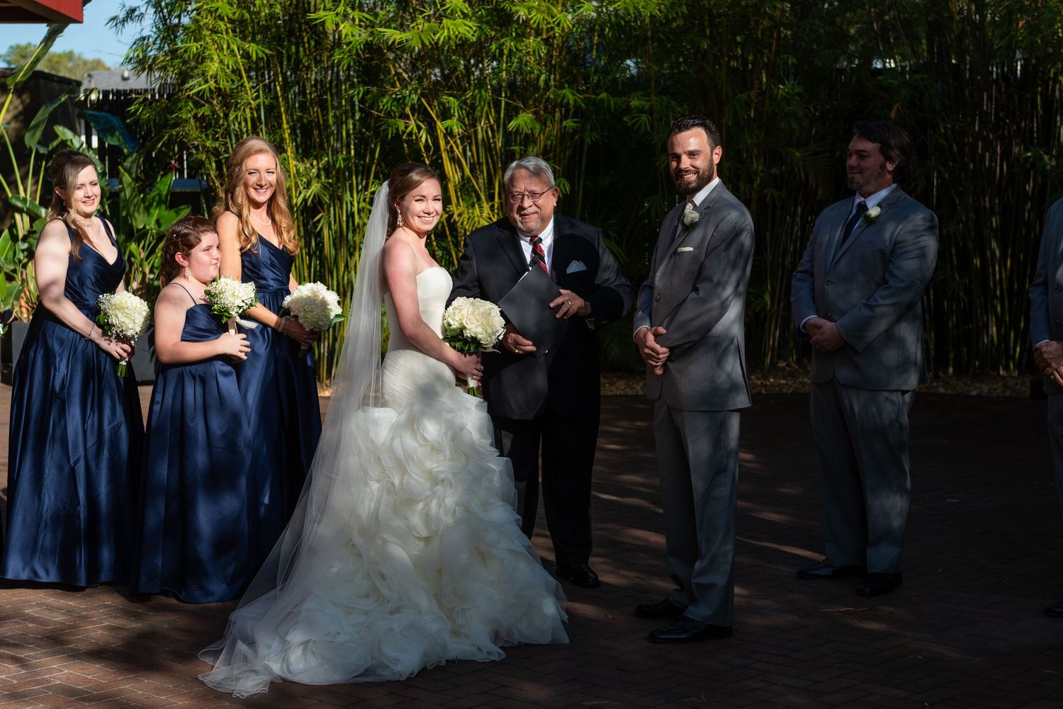 Wedding party poses outdoors with bride in white gown, bridesmaids in navy dresses, and groomsmen in gray suits.