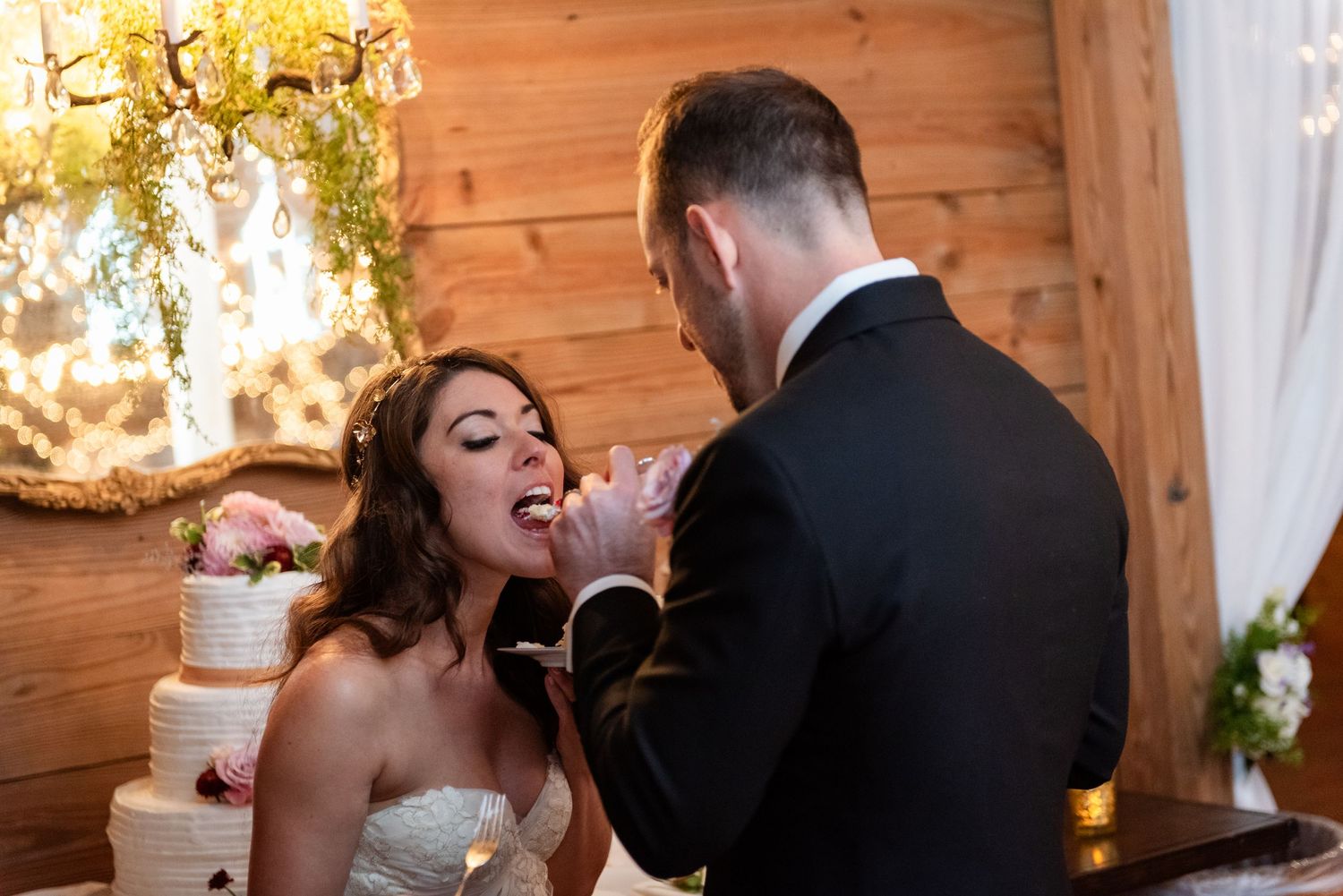Groom feeding wedding cake to his bride in a strapless gown near a multi-tier cake display.
