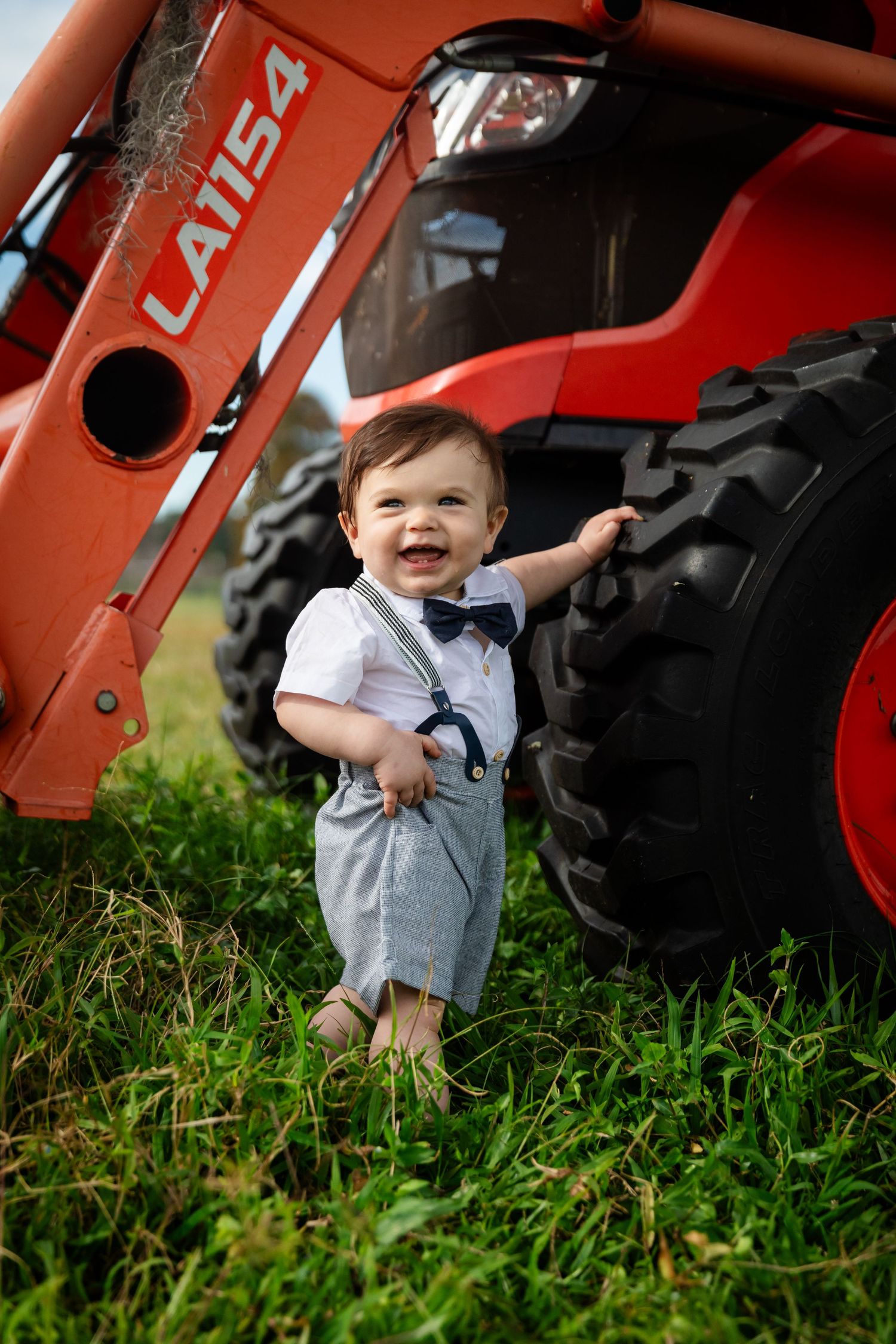Smiling baby boy in suspenders and bow tie standing by a red tractor on green grass.