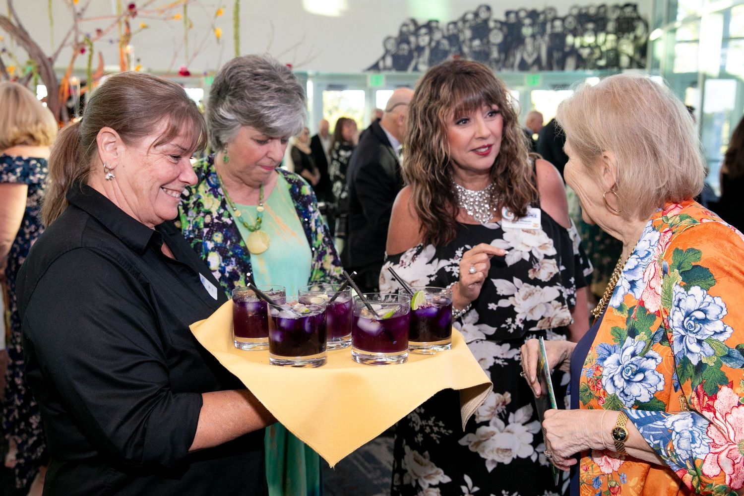 Women socializing at an elegant event while a server offers purple cocktail drinks on a tray.