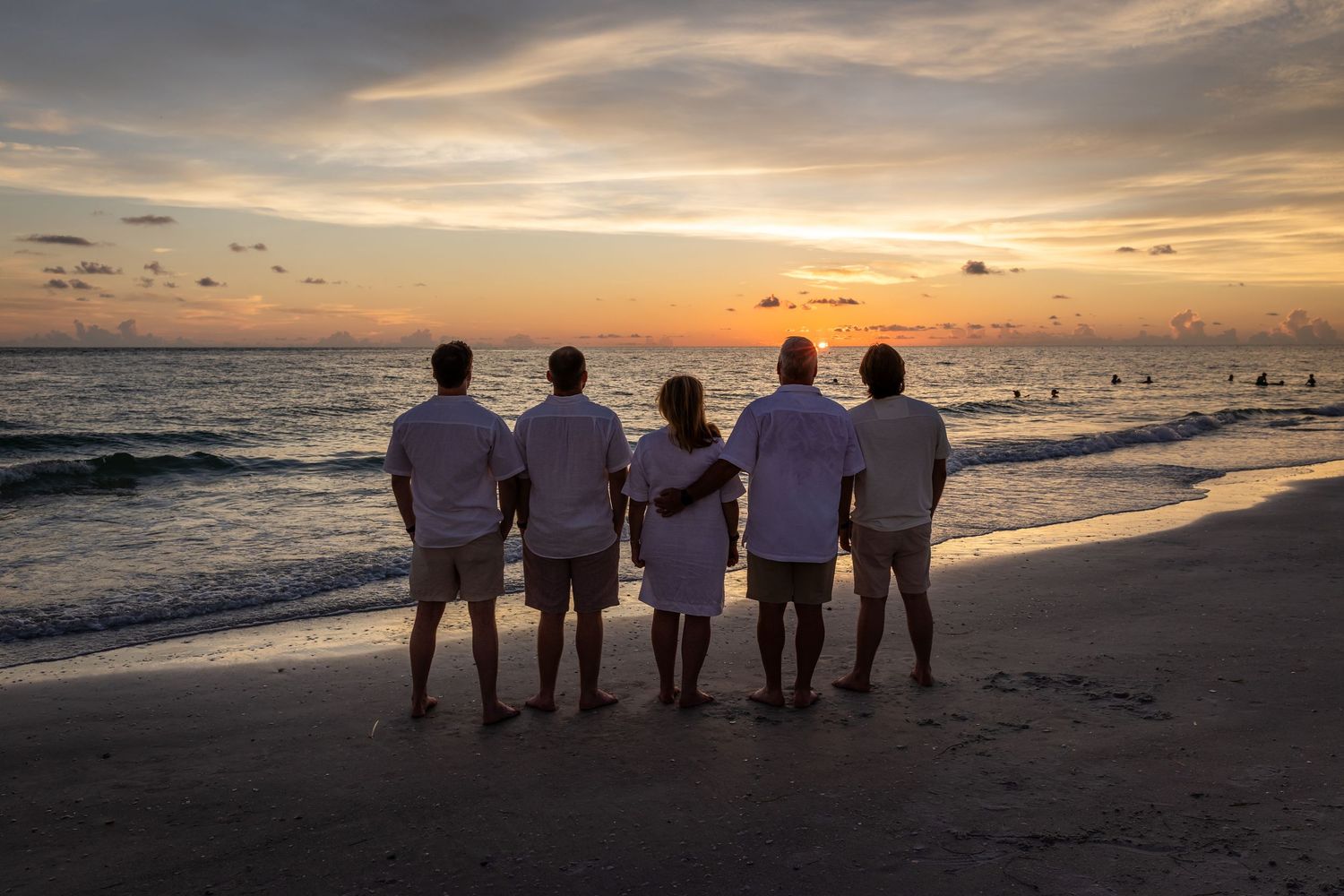 Five people in white shirts stand together on a beach watching a vibrant sunset over the ocean.