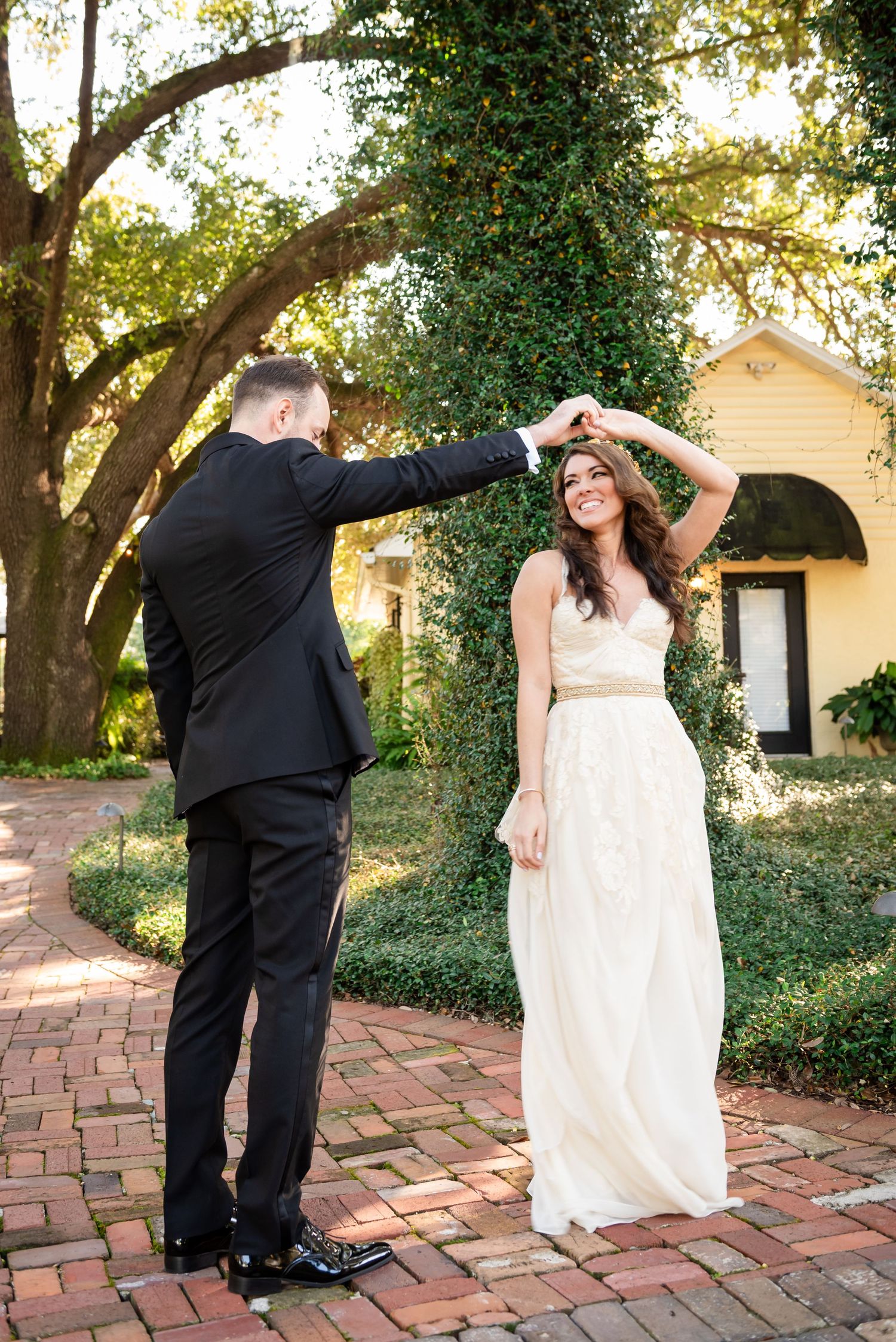 Groom twirls smiling bride in lace wedding dress on brick pathway surrounded by lush green trees.