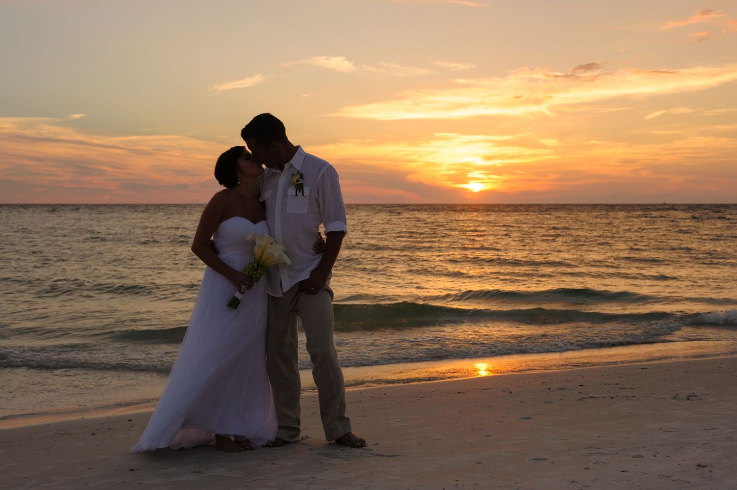 Newlywed couple kissing in silhouette on the beach during a breathtaking golden sunset.