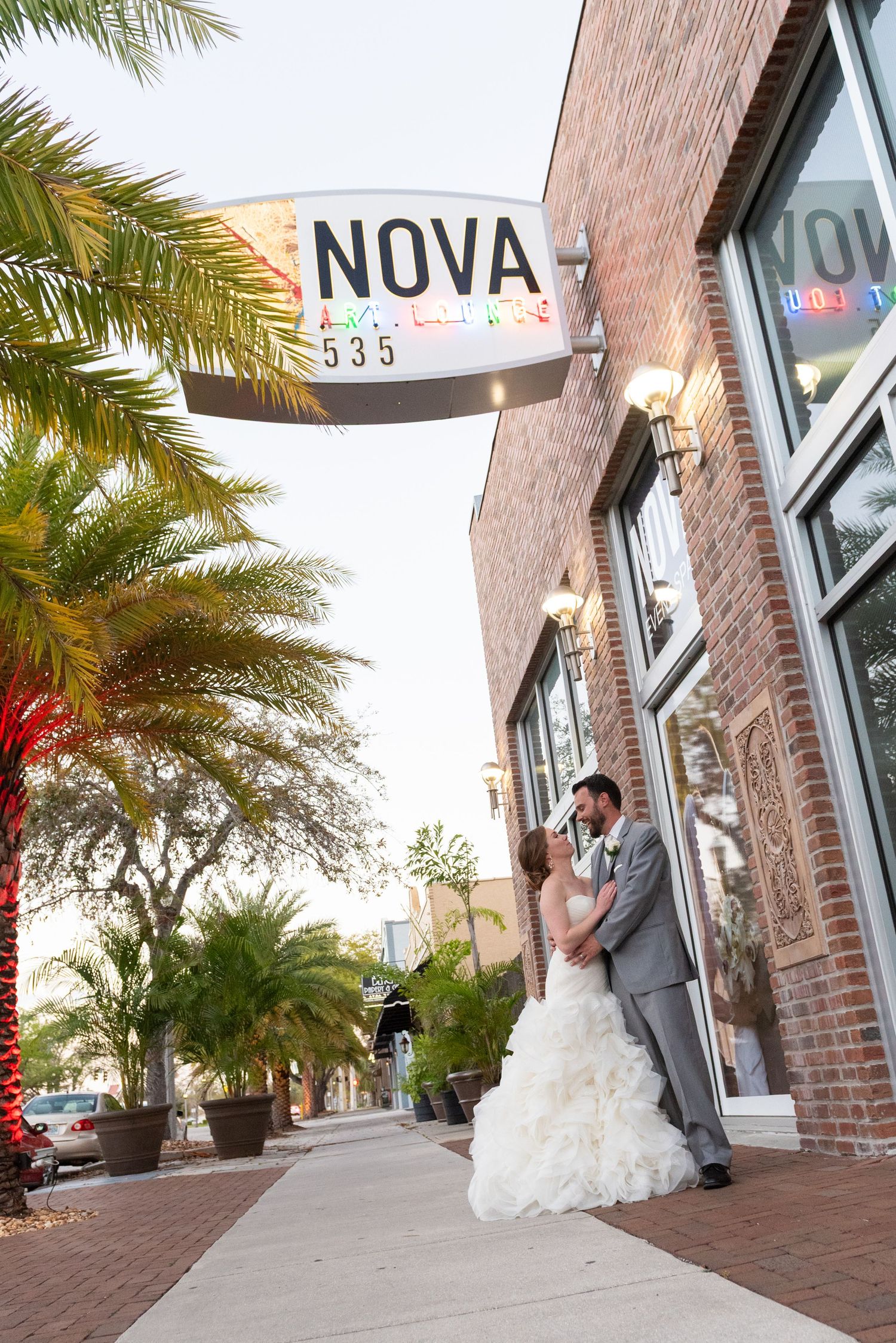 Bride and groom share romantic kiss under Nova 535 venue sign on brick building sidewalk.