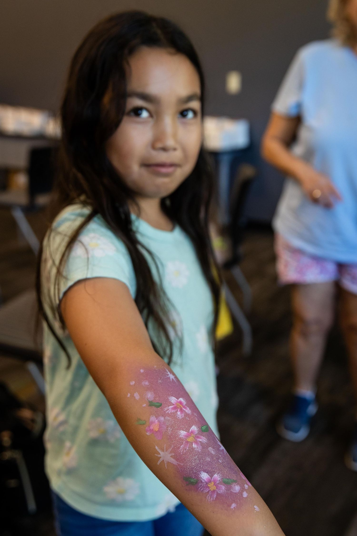 A young girl proudly displays colorful floral arm painting with pink flowers at an indoor event.