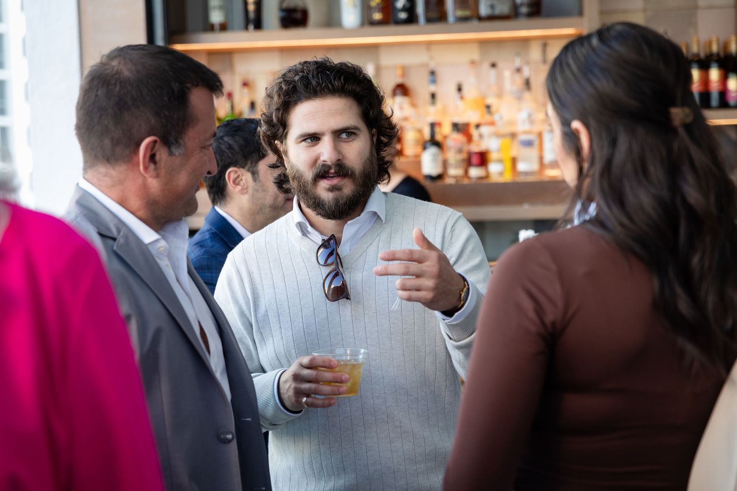 Man holding a drink conversing with guests at a bar social event in a white sweater with sunglasses.