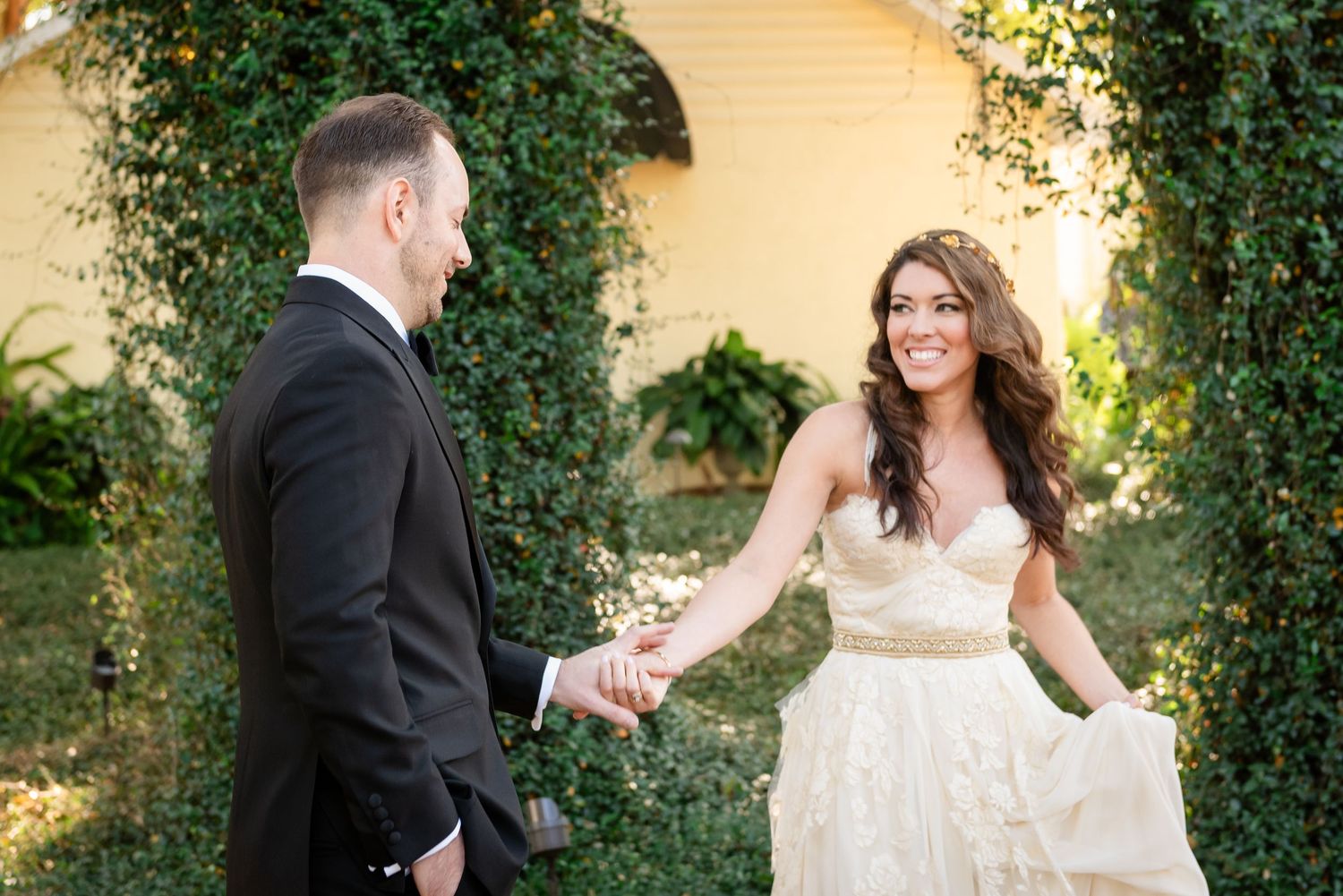 Bride in strapless lace gown holds hands with groom in black tuxedo outdoors near ivy-covered wall.
