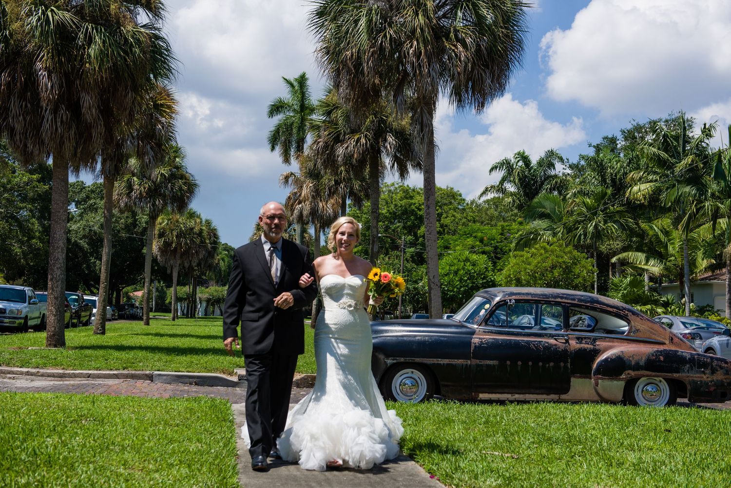 Bride and groom pose outdoors near vintage car with palm trees on sunny wedding day in Florida.